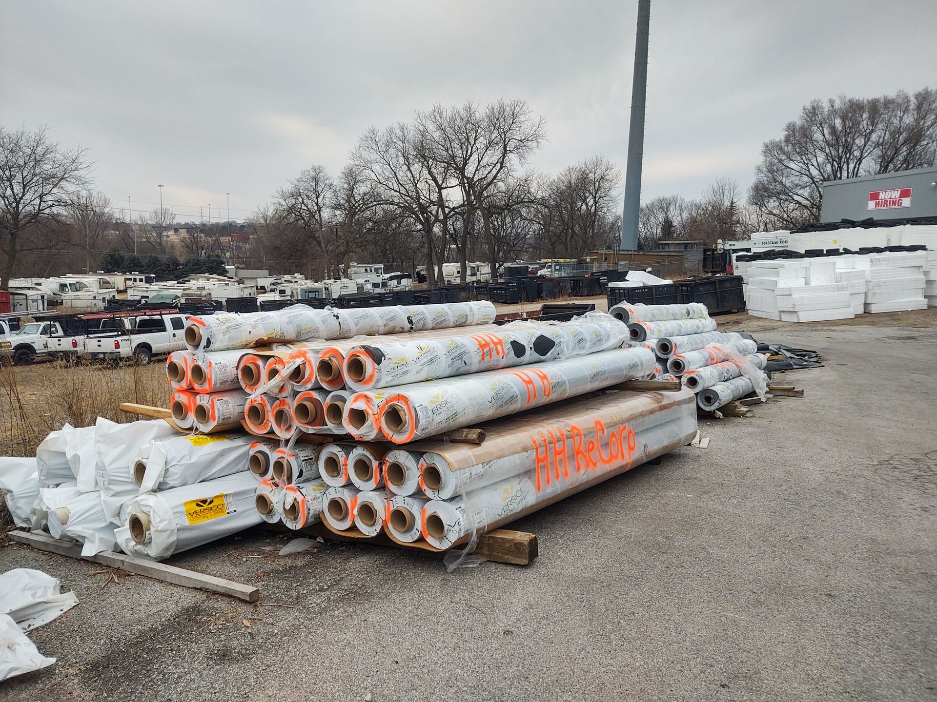 Stacked rolls of construction material with orange labels sit on a gravel lot near a parked truck and building materials.