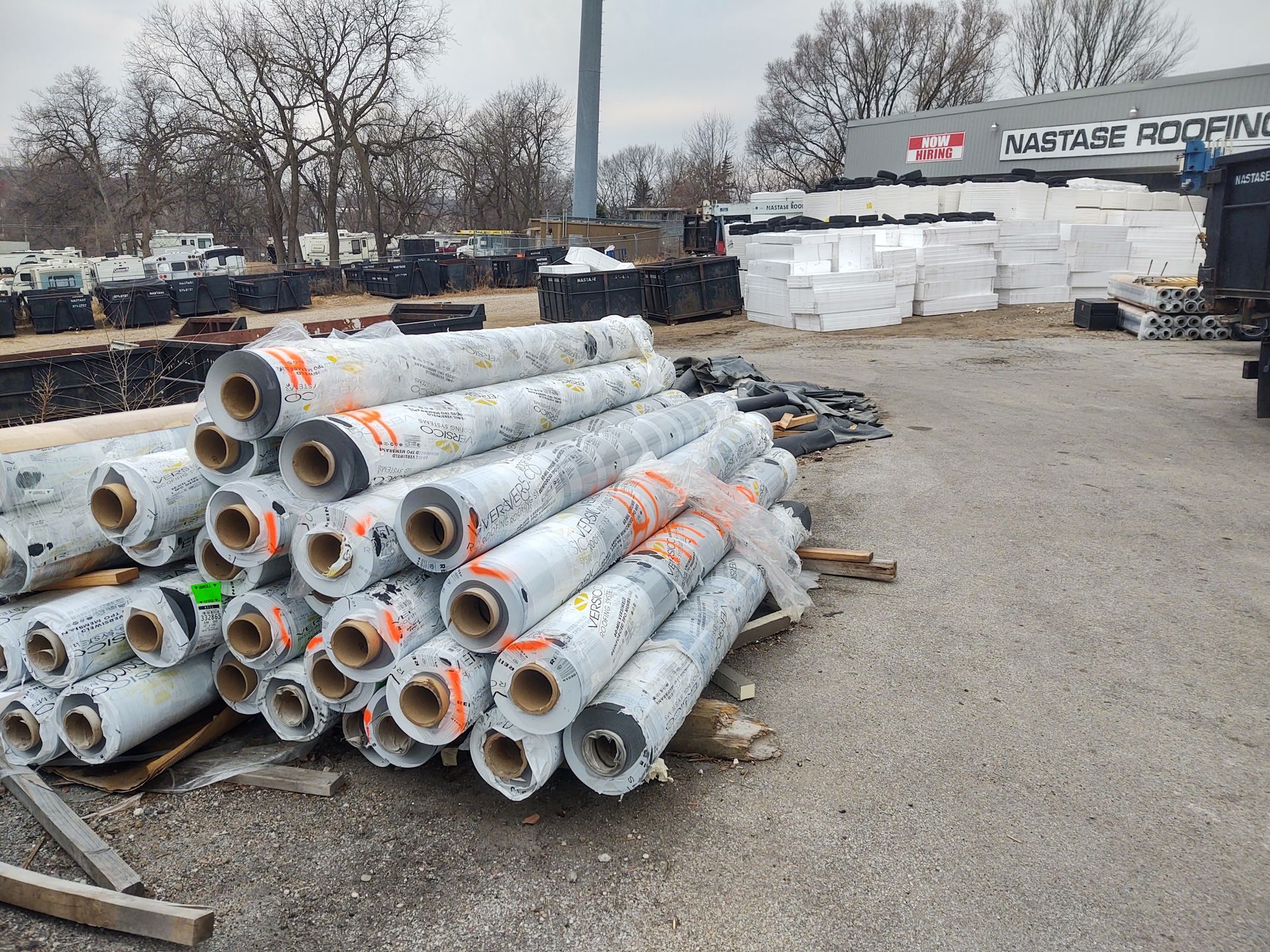 A large pile of shrink-wrapped building material rolls sits on a gravel lot outside a roofing supply business.