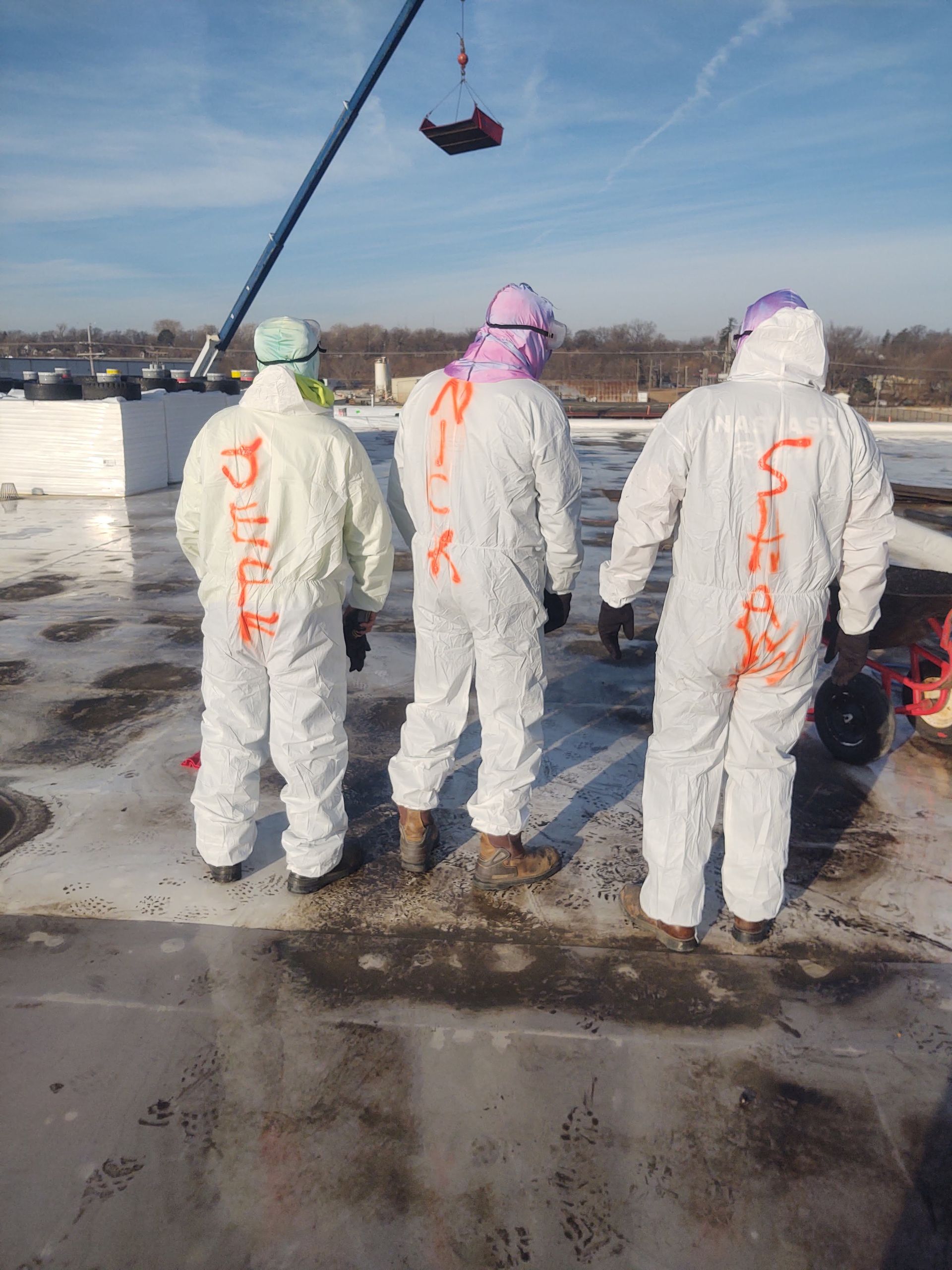 Three workers in white protective suits with names written on the back stand on a flat roof under a clear blue sky.