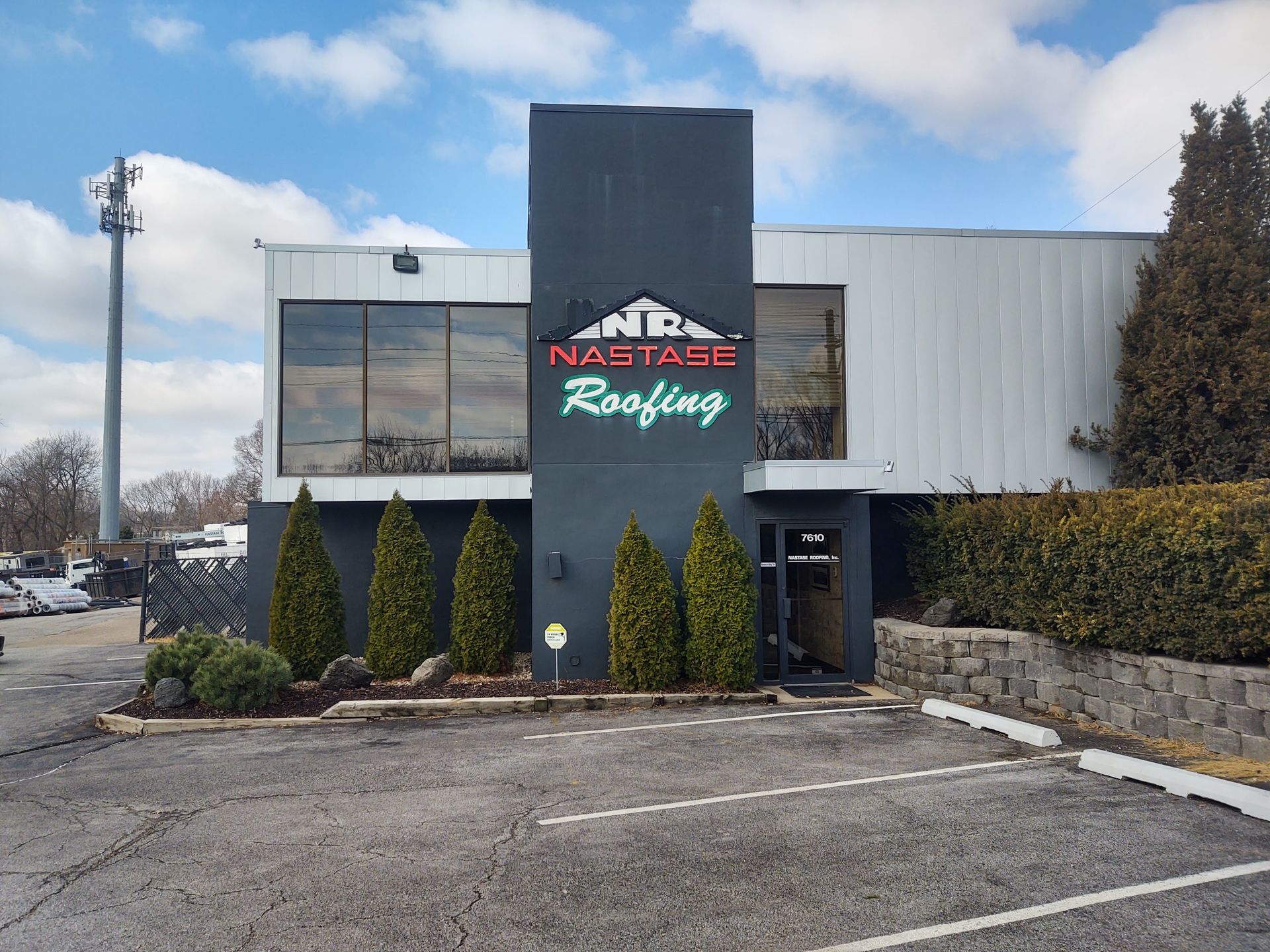 The Nastase Roofing building features a dark gray central column with signage, flanked by white walls and small evergreens.