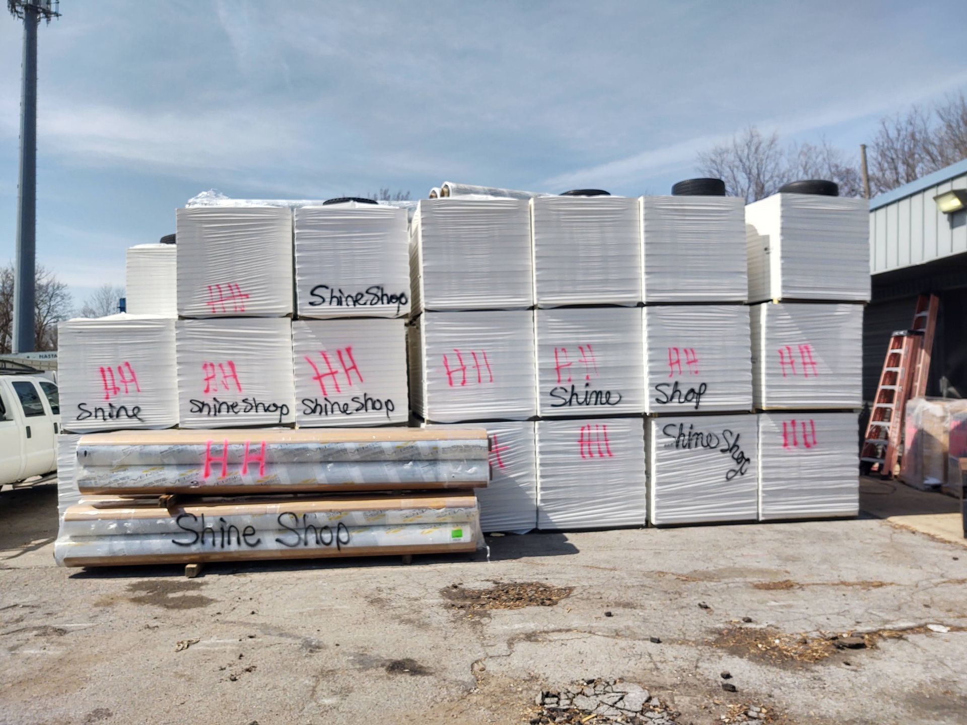 Stacked pallets of white, shrink-wrapped material marked with red lettering in an outdoor lot near a building.