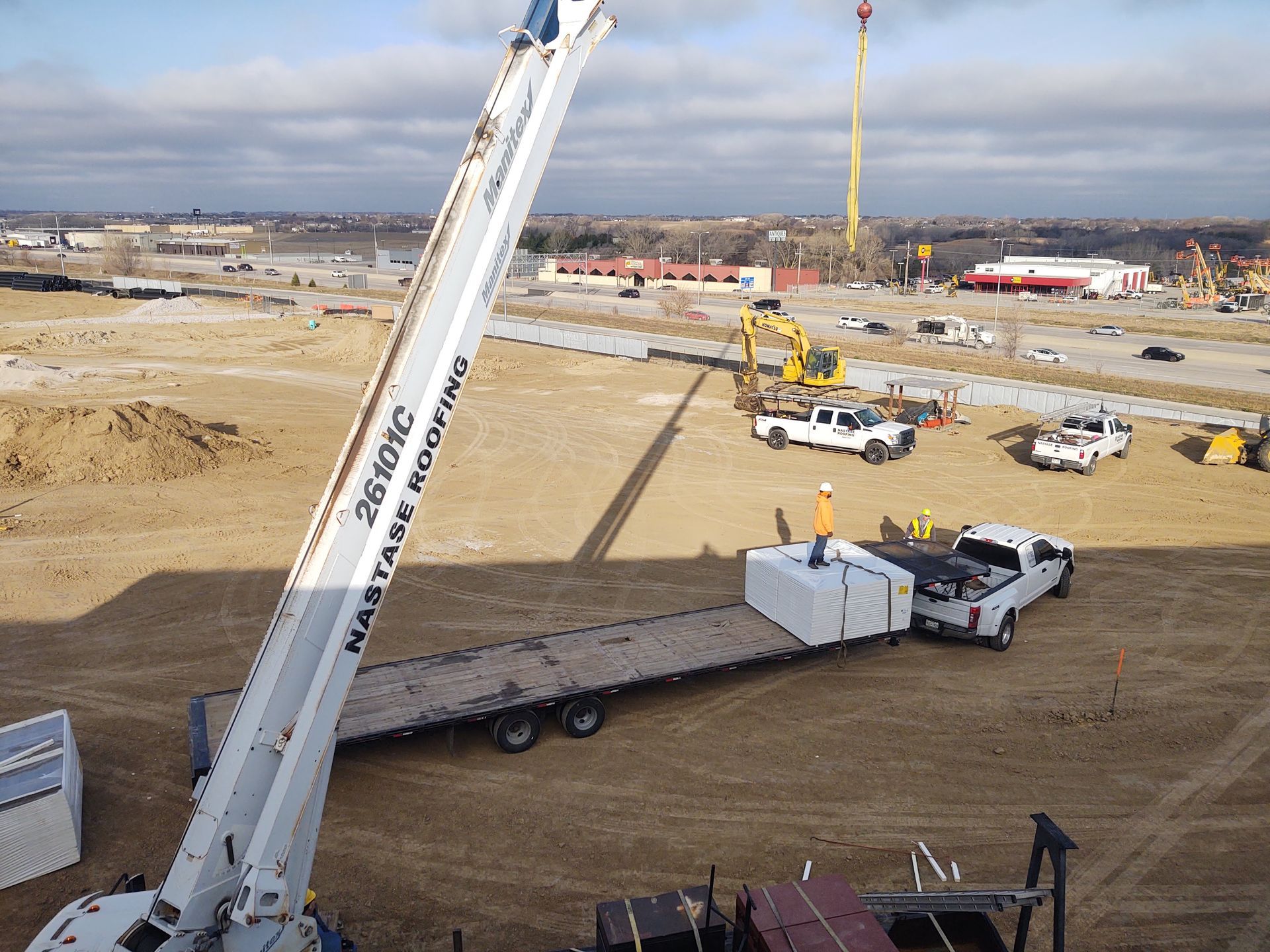 A crane and flatbed trailer with a white load parked on a construction site, with heavy equipment and a road in the back.