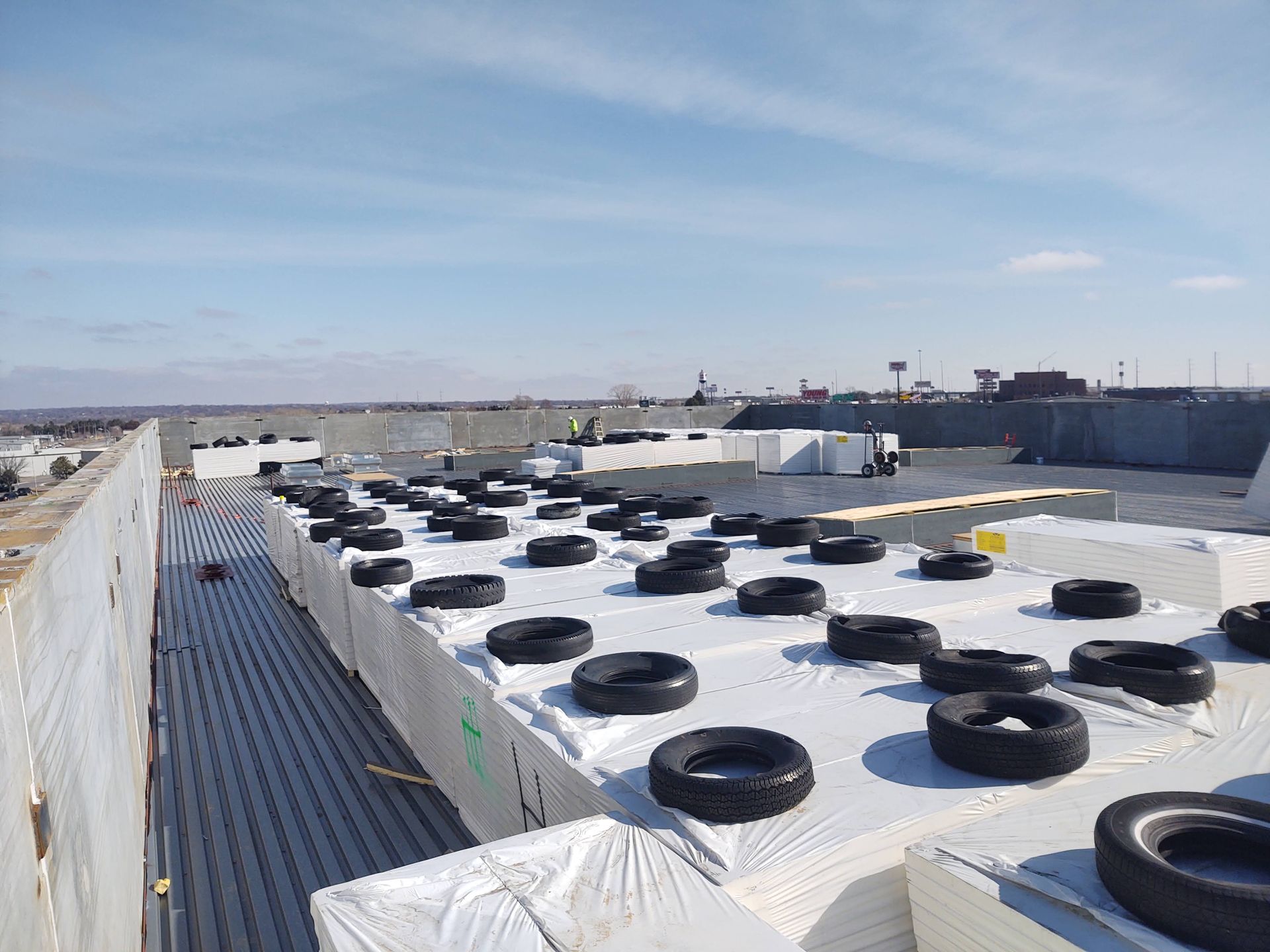 Construction workers use old tires to weigh down white insulation panels on a flat, sunny rooftop.