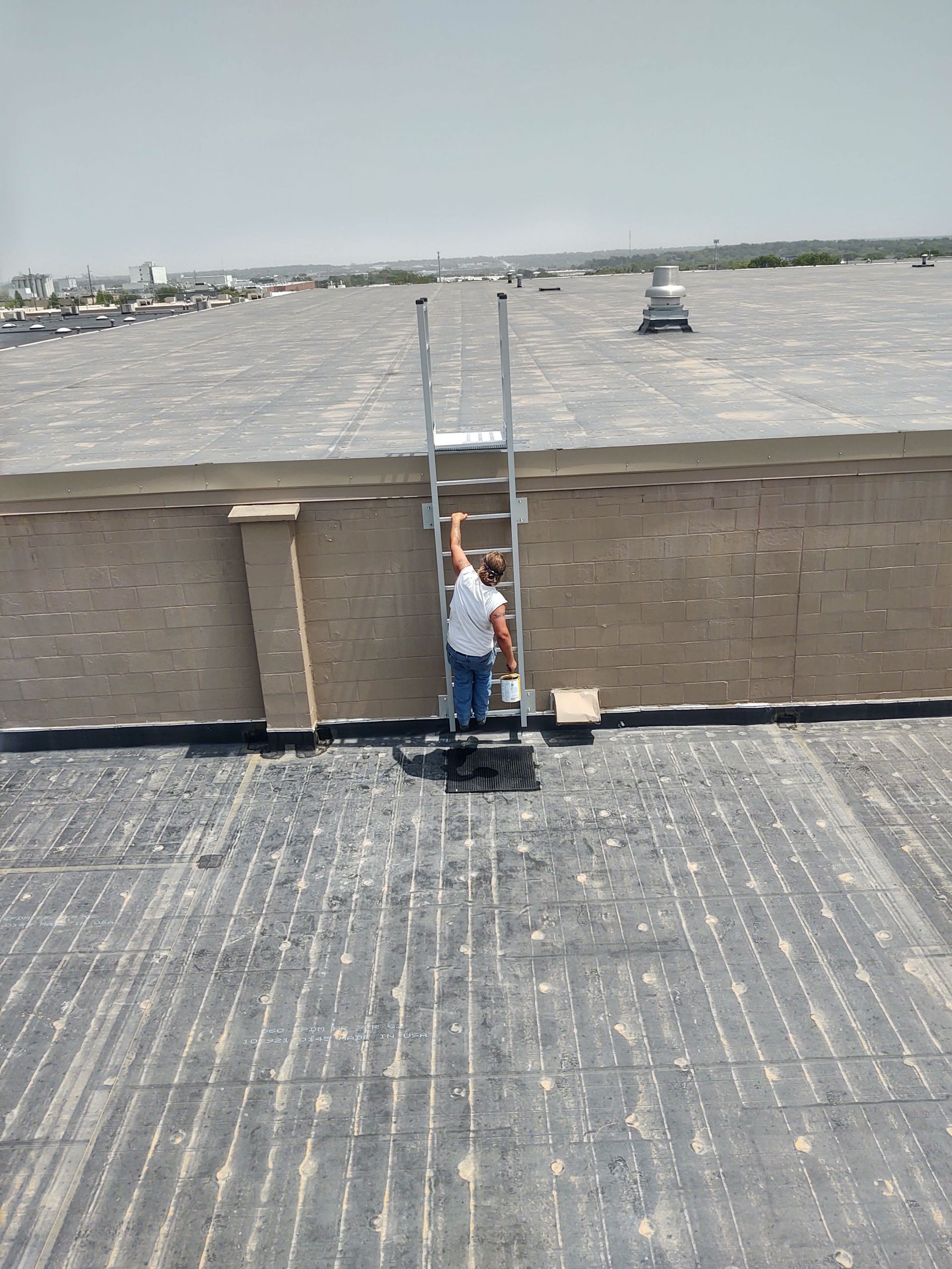 A worker in a white shirt climbs a metal ladder leaning against a brick parapet wall on a flat, gravel-covered roof.