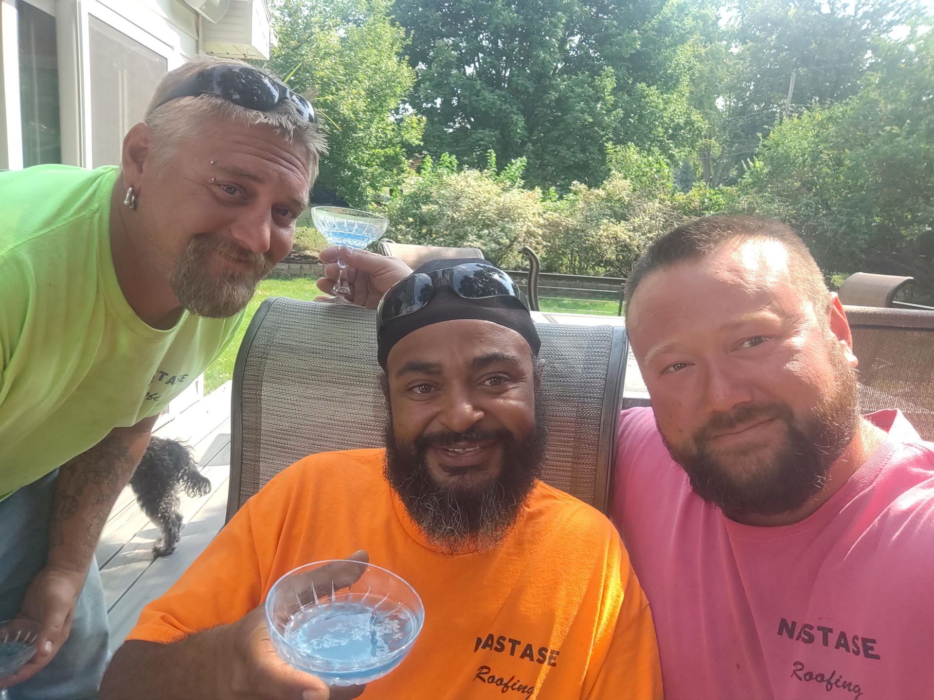 Three men in colorful shirts pose together outdoors, smiling and holding drinks in a bright, sunlit setting.