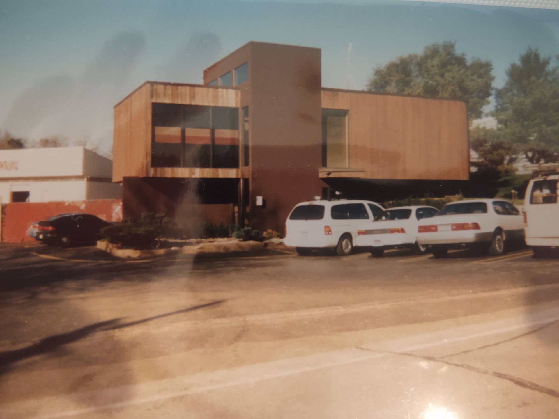 A two-story brown building with wood paneling and large windows, situated above a parking lot with several parked cars.