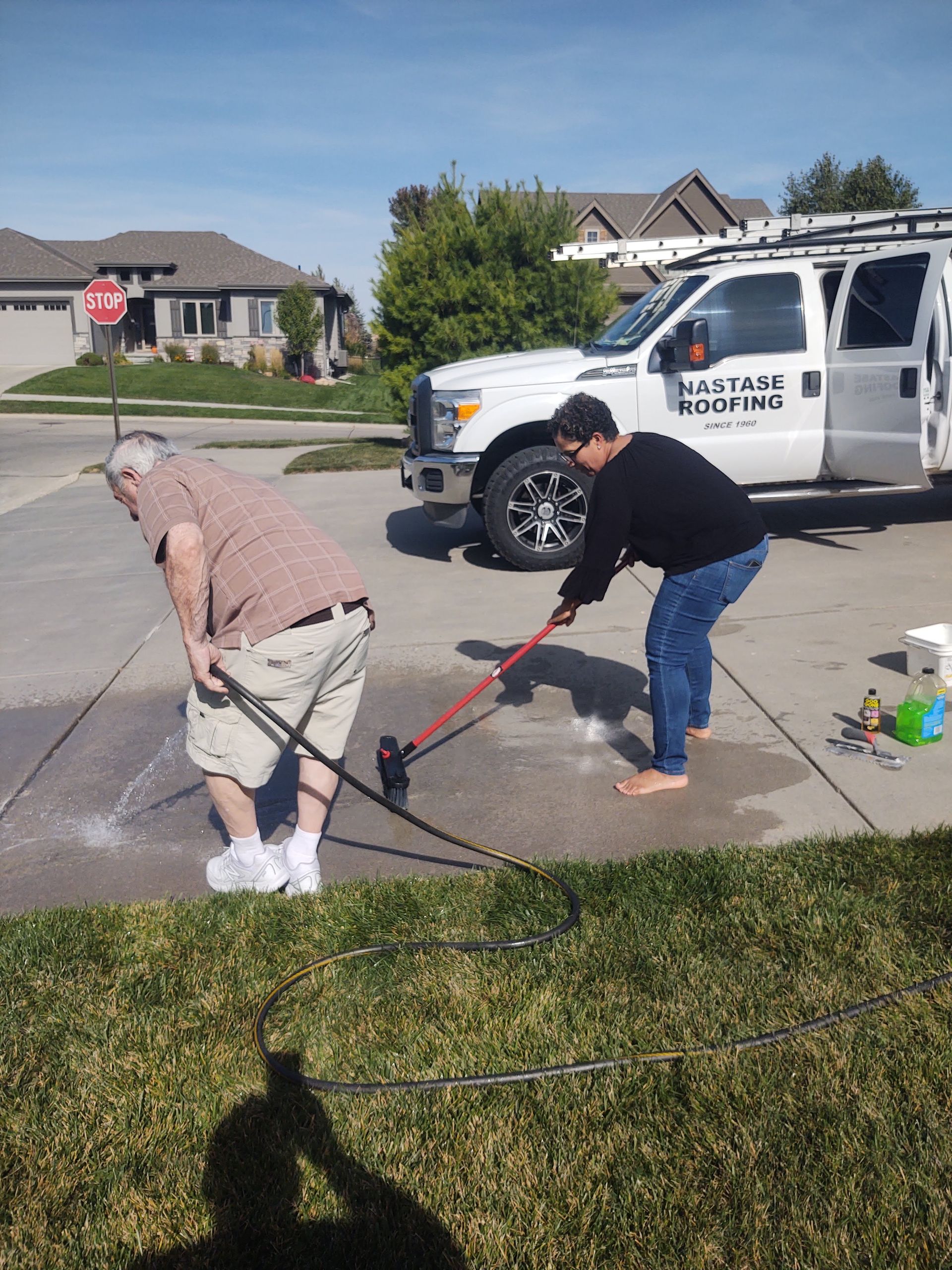 Two people clean a concrete driveway with a pressure washer, standing near a white service van on a sunny day.