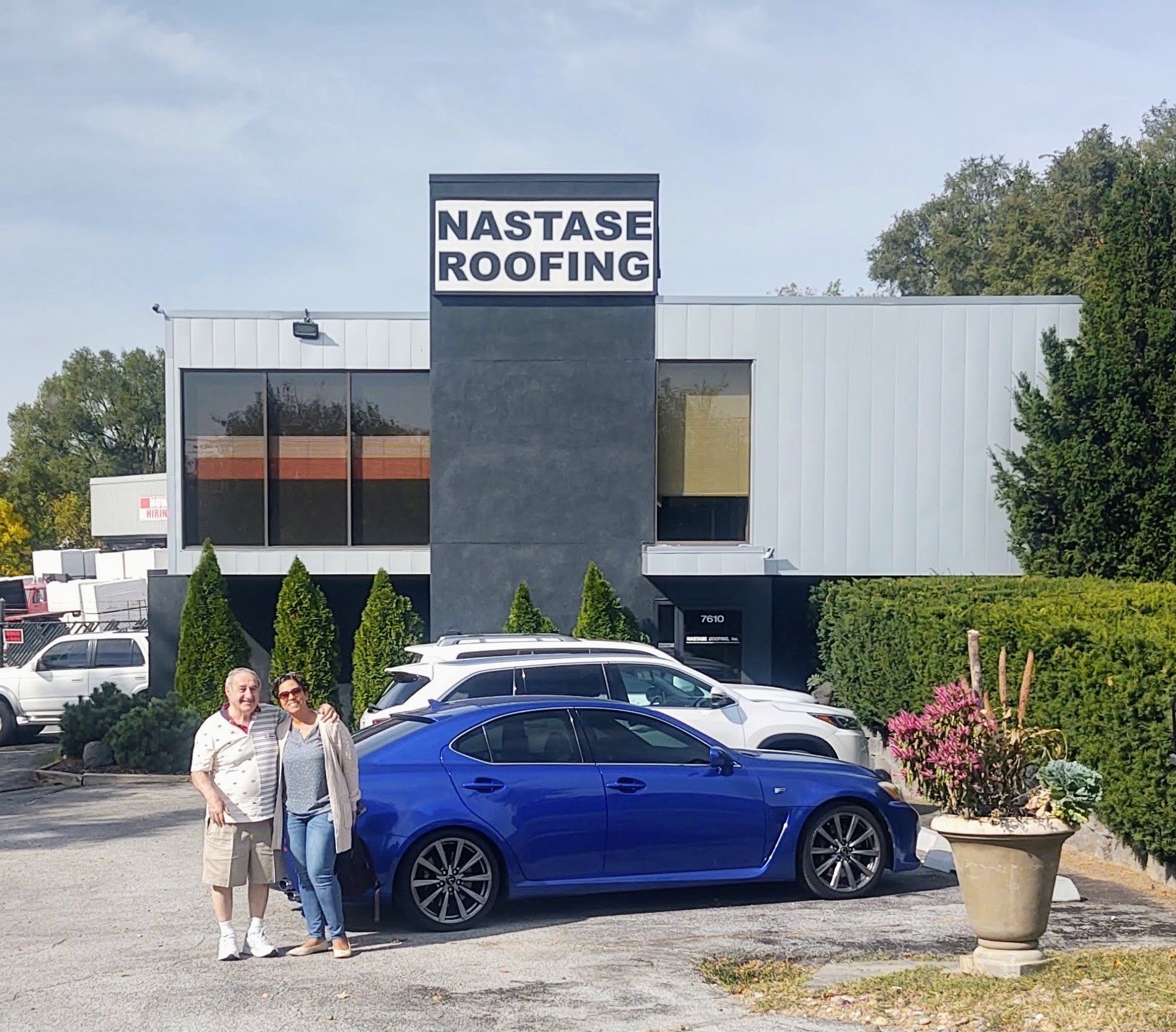 A couple poses in front of a blue sedan parked at the Nastase Roofing office building on a sunny day.