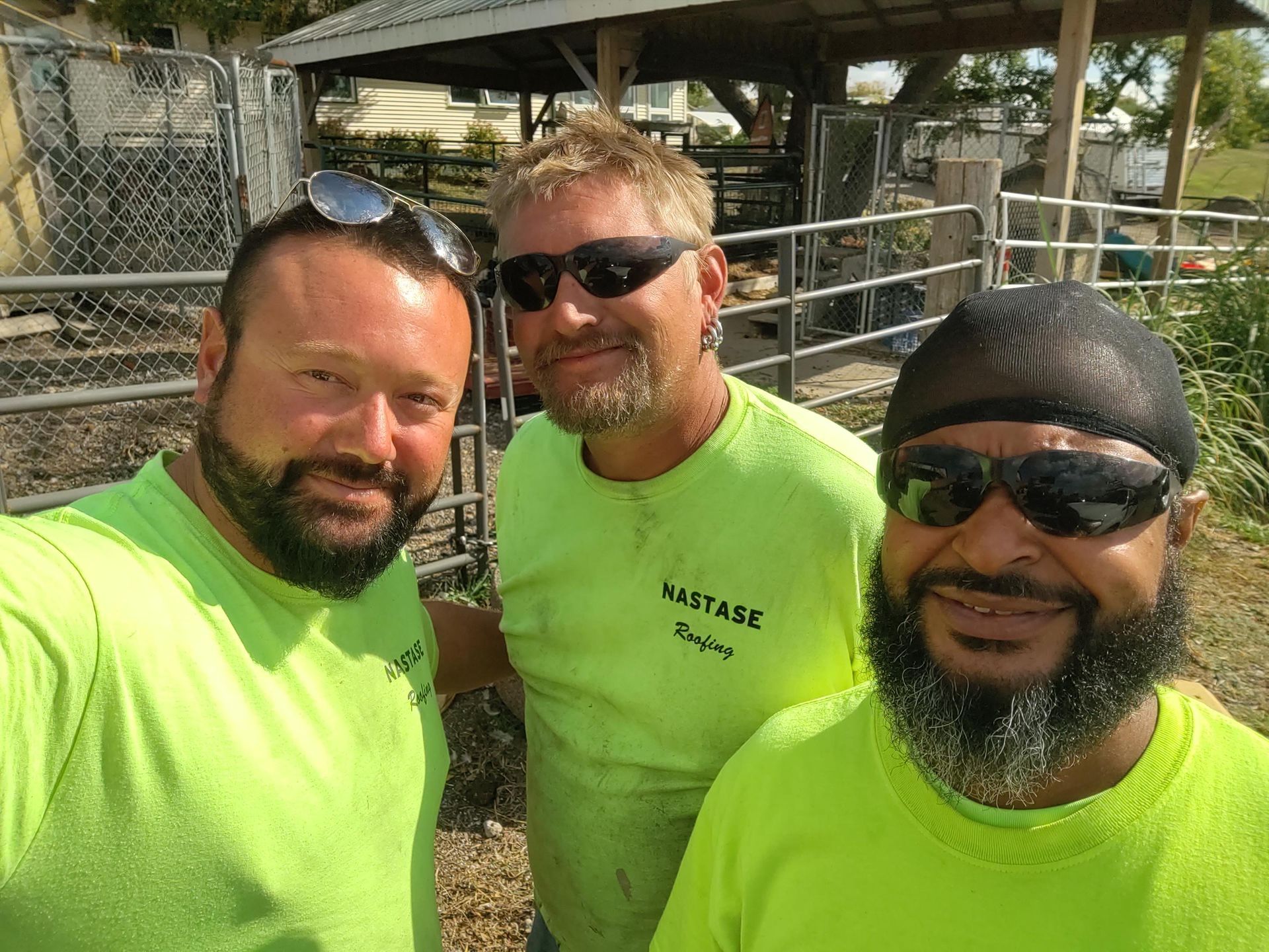 Three people wearing neon yellow shirts and sunglasses stand outdoors in front of a metal fence and structure.