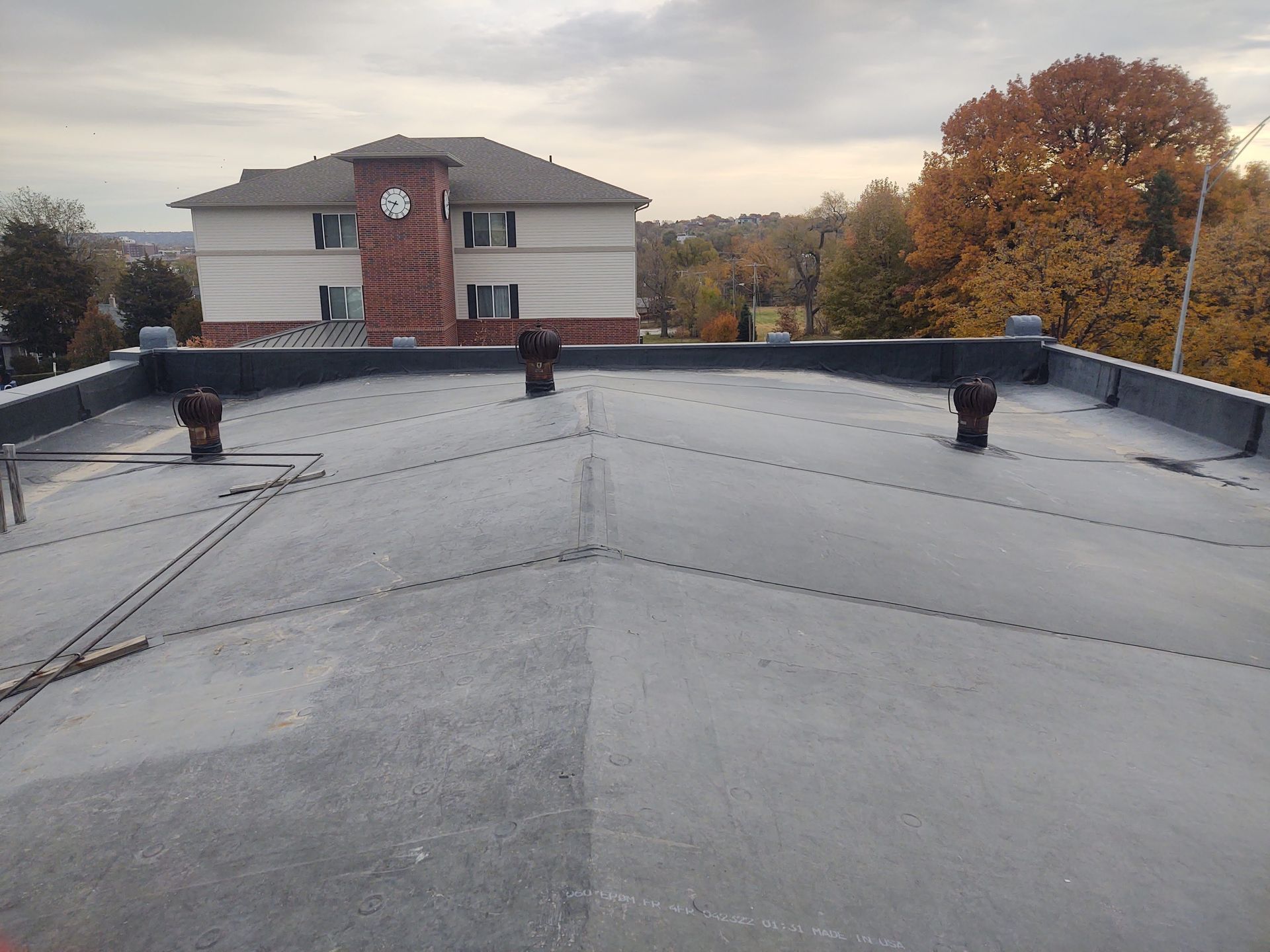 A flat grey roof with three vents, seen from a high angle, with a building and autumn trees in the background.