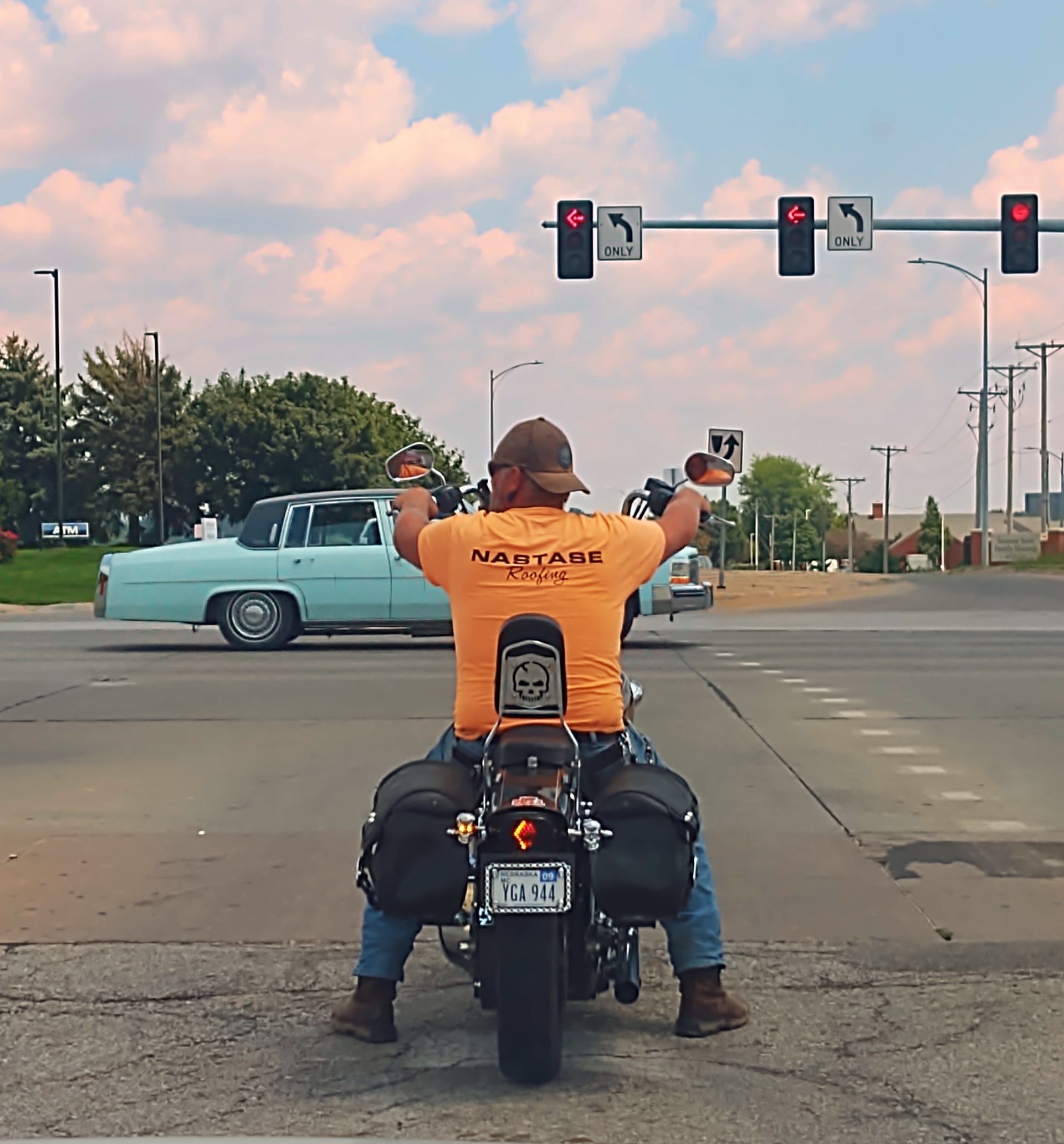 A motorcyclist in an orange shirt sits on a motorcycle at an intersection, with a light blue classic car driving by.