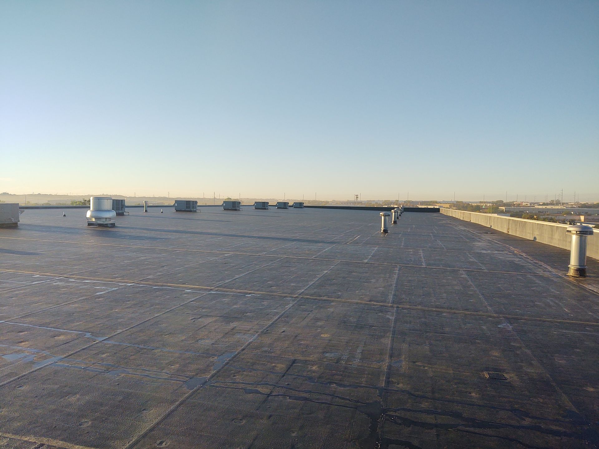 A flat, dark, gravel-covered commercial roof under a clear blue sky, featuring several small ventilation pipes.