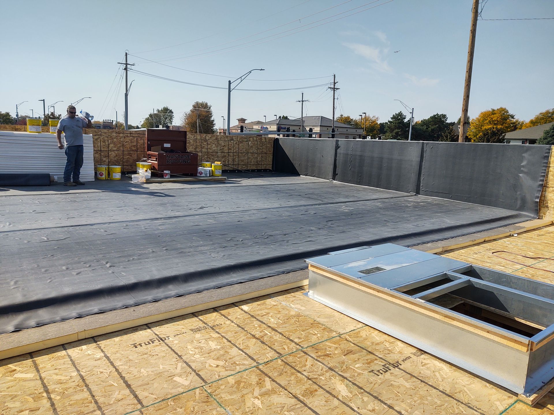 A construction worker stands on a rooftop deck with a partially installed black membrane and a metal roof curb.