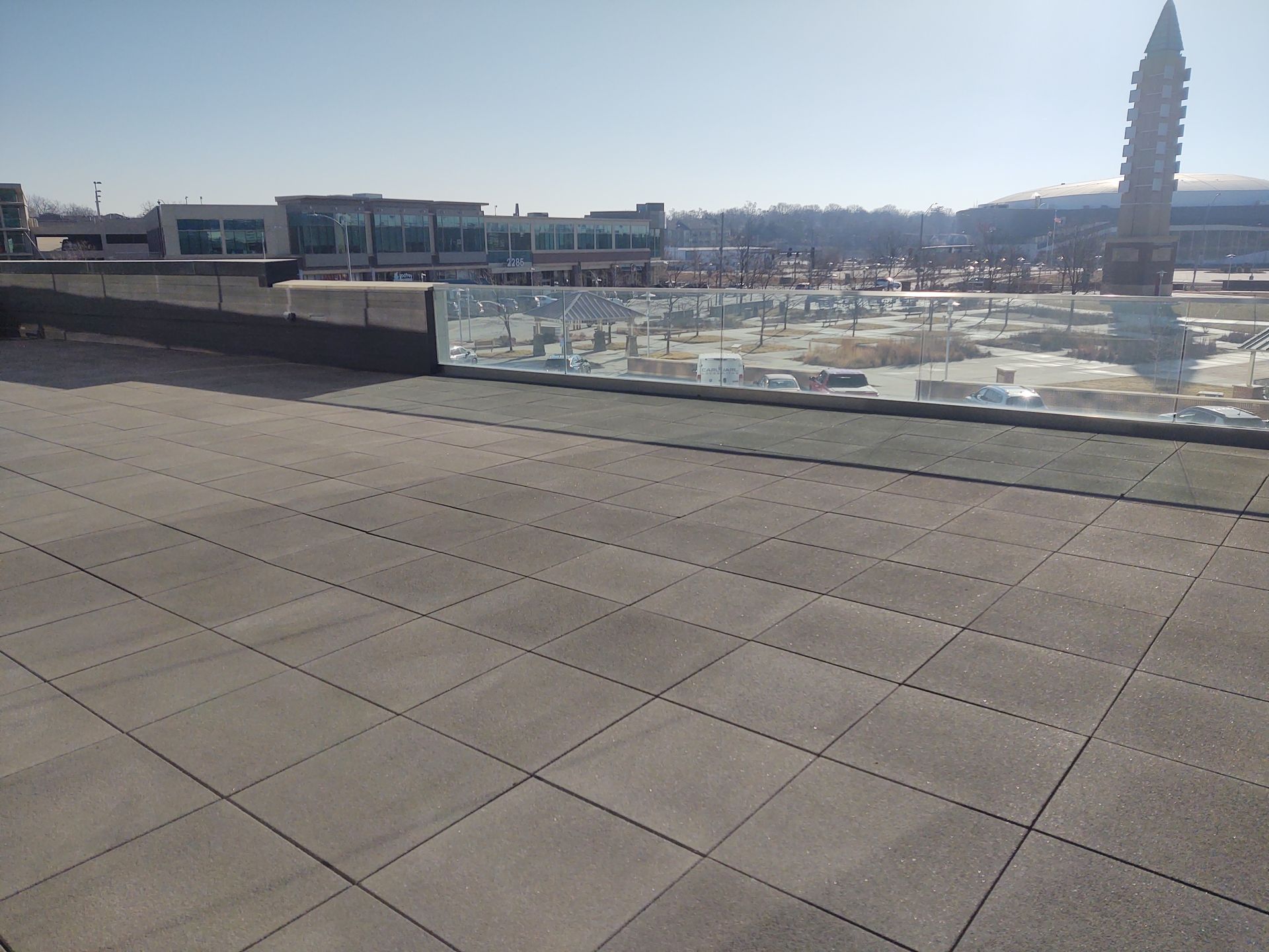A paved terrace with a glass railing overlooks a plaza featuring a tall, slender sculpture under a clear blue sky.