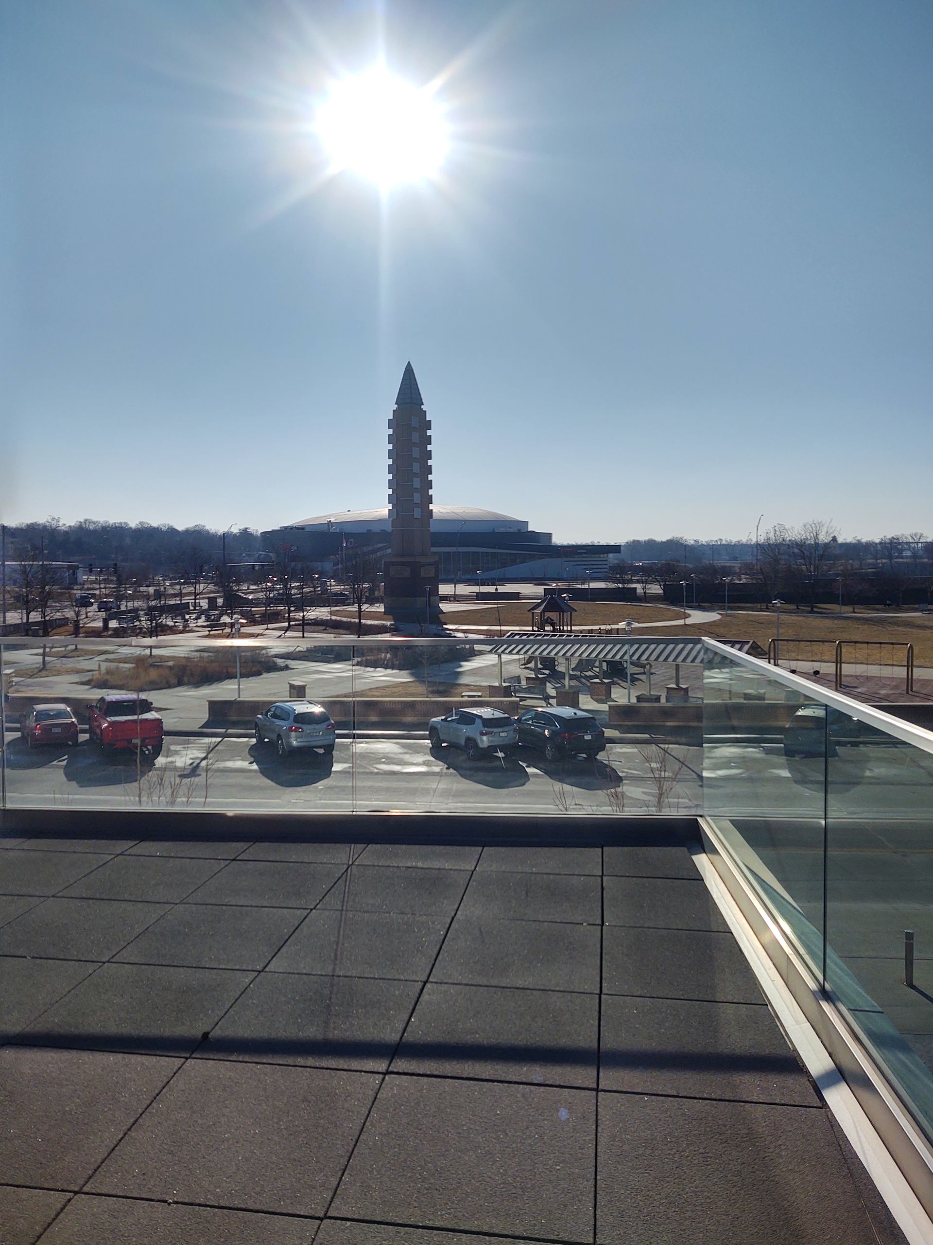 A tall, stone obelisk stands in a sunny parking lot near a low building under a clear blue sky.