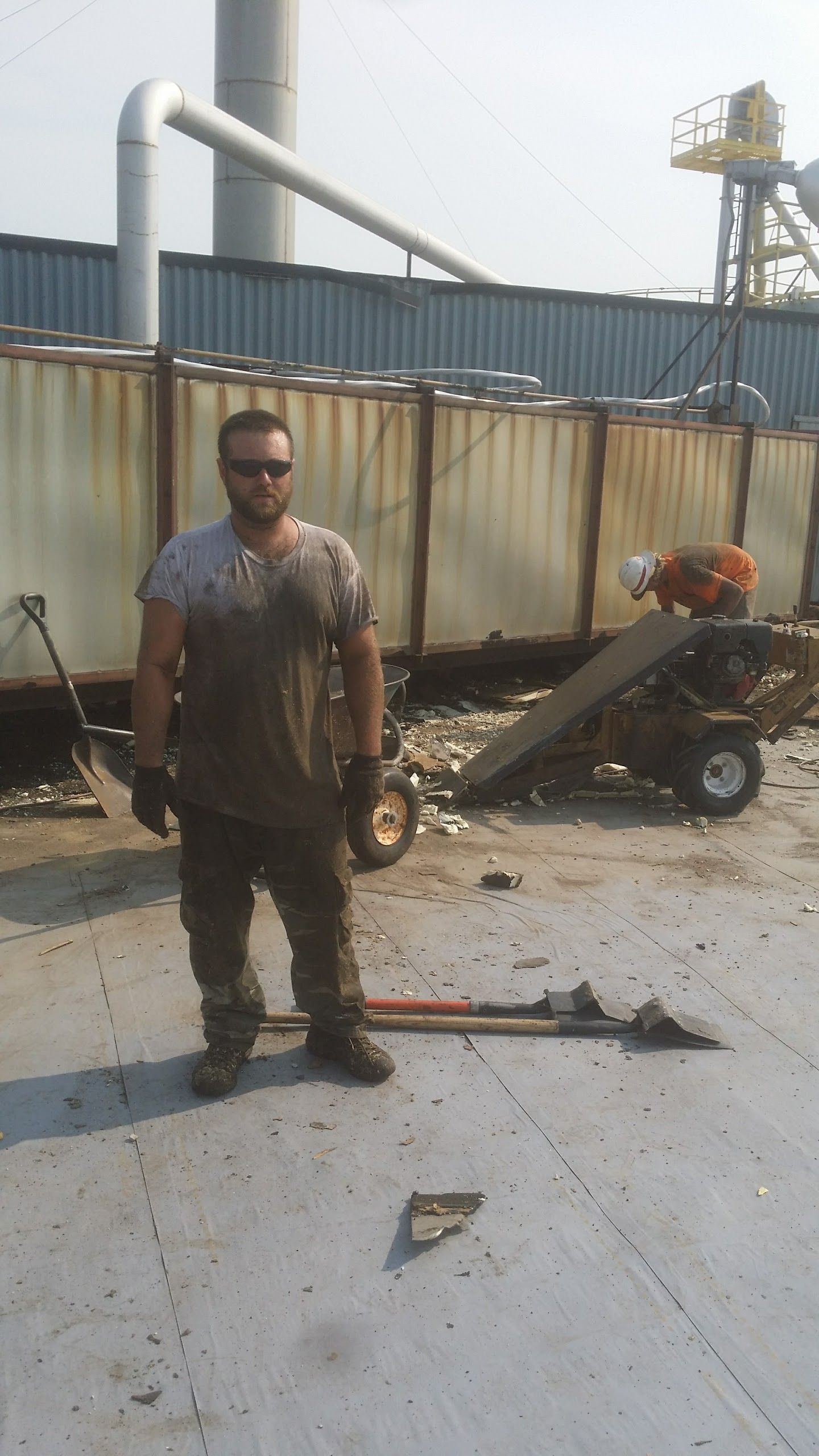 A person wearing work clothes and sunglasses stands on a construction site next to a dumpster and equipment.
