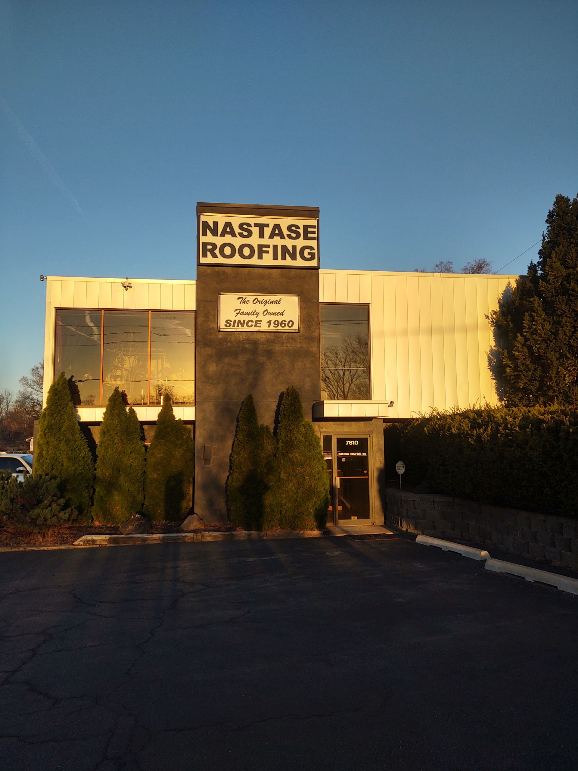 The Nastase Roofing commercial building at dusk with a prominent sign, dark pillar, and surrounding evergreen bushes.