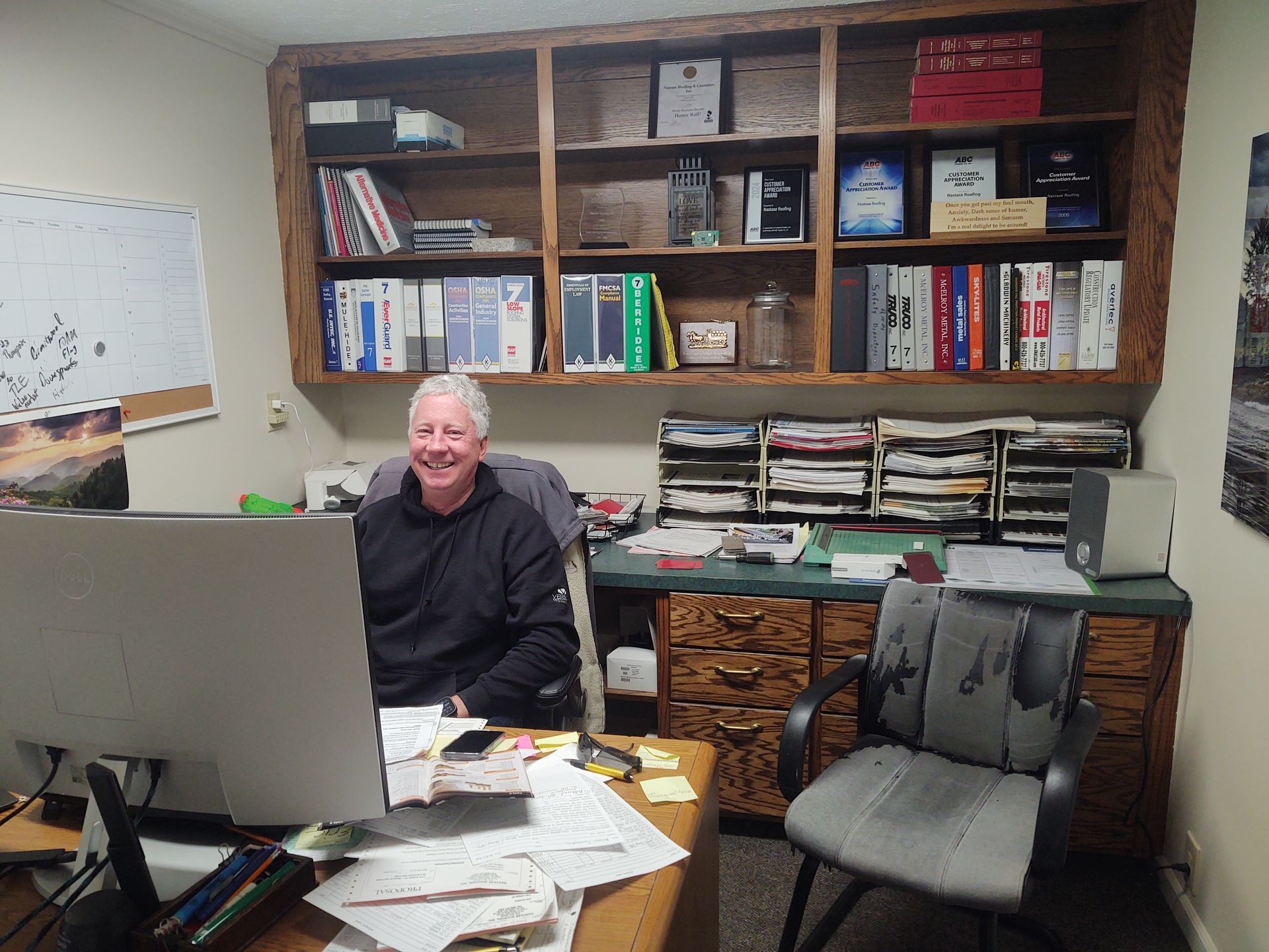 A smiling person sits at a cluttered desk in an office with wooden bookshelves filled with binders and files.