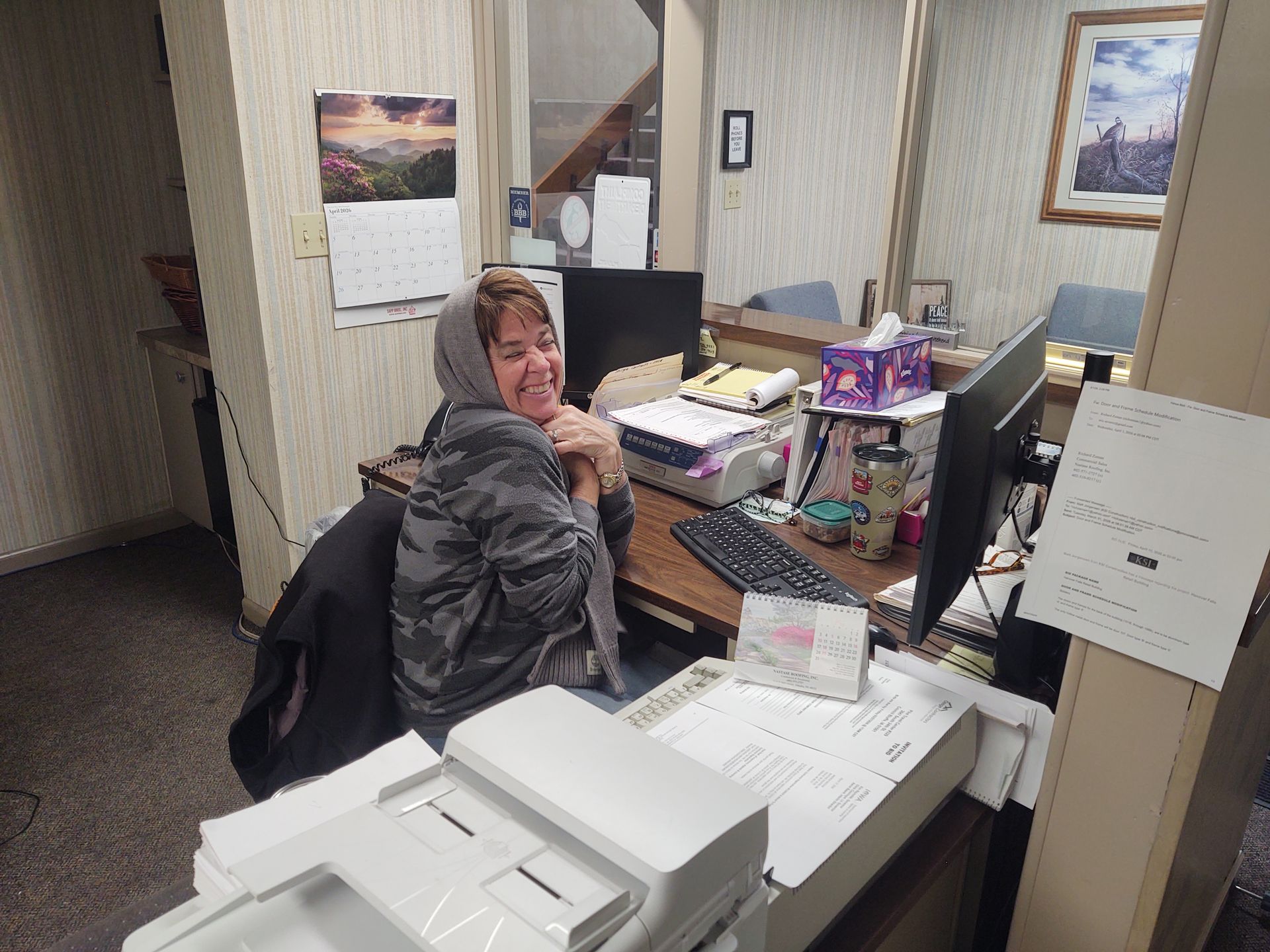 A person wearing a gray hoodie sits at a cluttered office desk, smiling at the camera while working on a computer.
