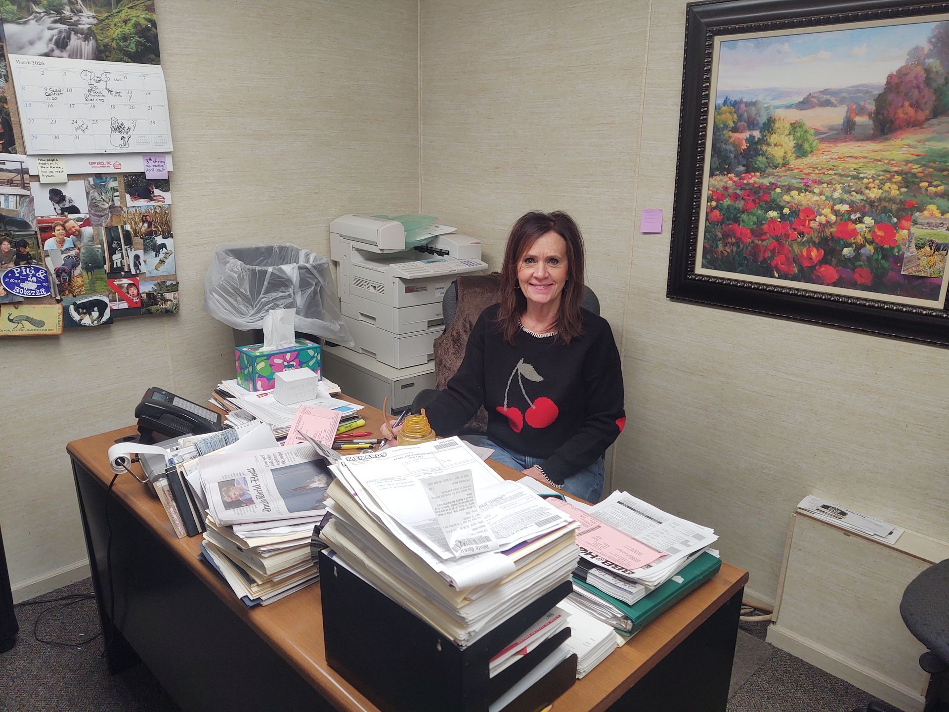 A smiling person sits at a cluttered office desk with paperwork and a printer behind them, next to a large landscape painting.