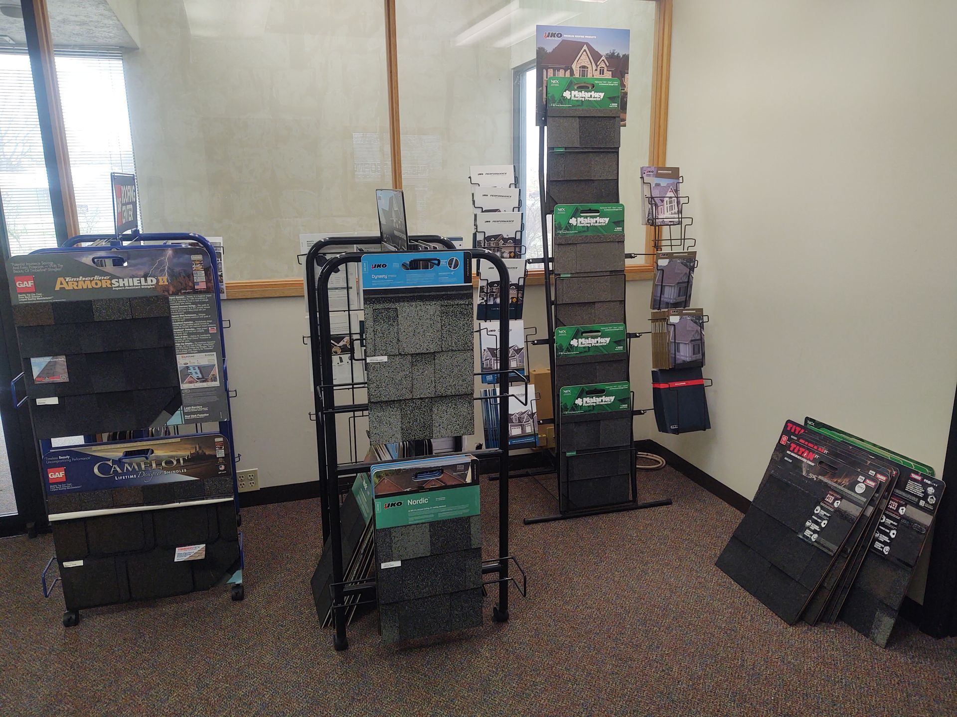 Display racks in an office showing various samples of dark-colored residential roofing shingles.