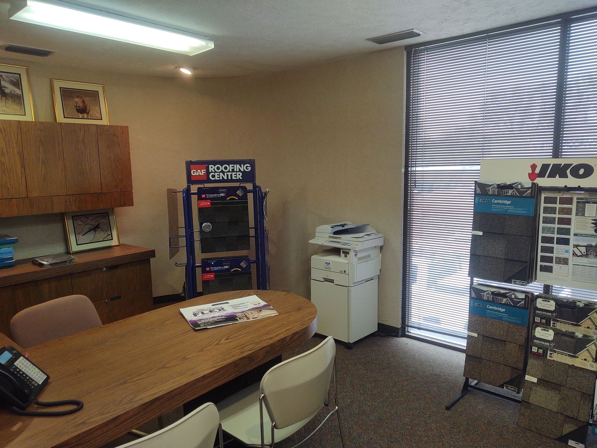 Office interior with a wooden conference table, office chairs, printer, cabinets, and two IKO product display racks.