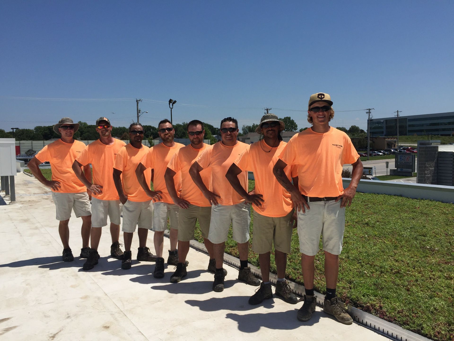 Eight people wearing matching orange shirts and khaki shorts stand in a row on a rooftop under a bright blue sky.