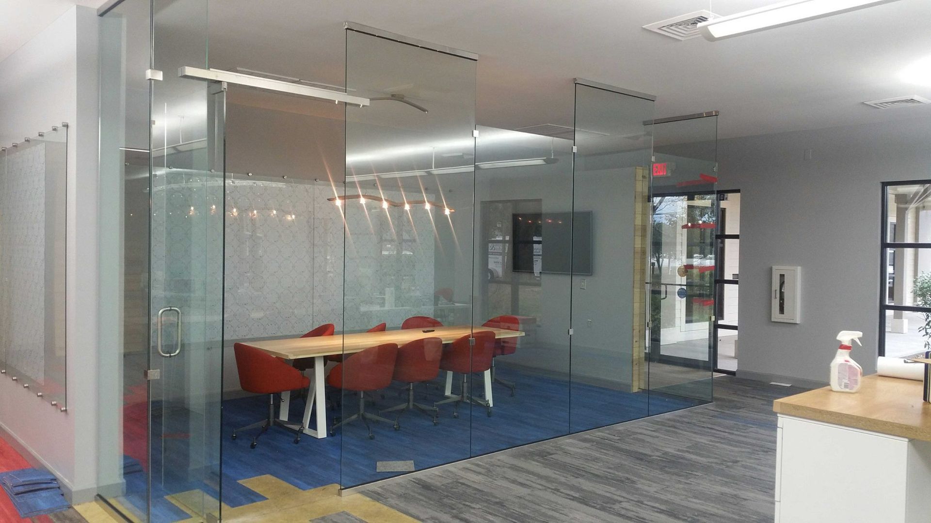 Modern office with glass walls, conference table, red chairs, blue carpet.