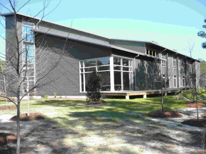 Gray modern building with large windows and sloped roof, set on a grassy area with trees.