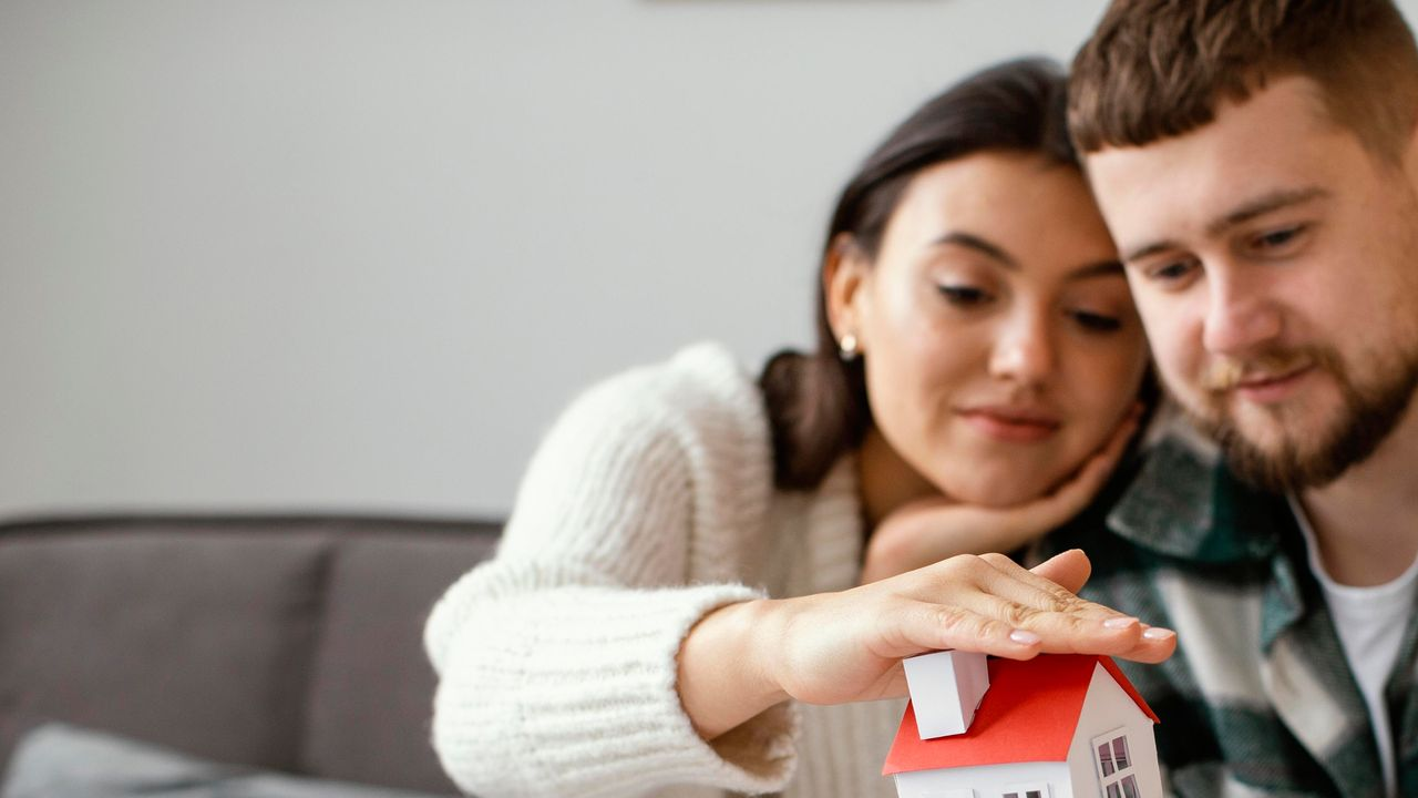 Couple looking at a miniature house, one protecting it, sitting on a sofa with paperwork.