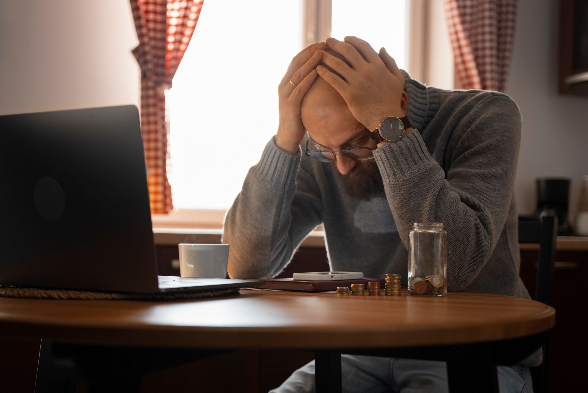 Man with hands on head, looking stressed at laptop and coins on table.