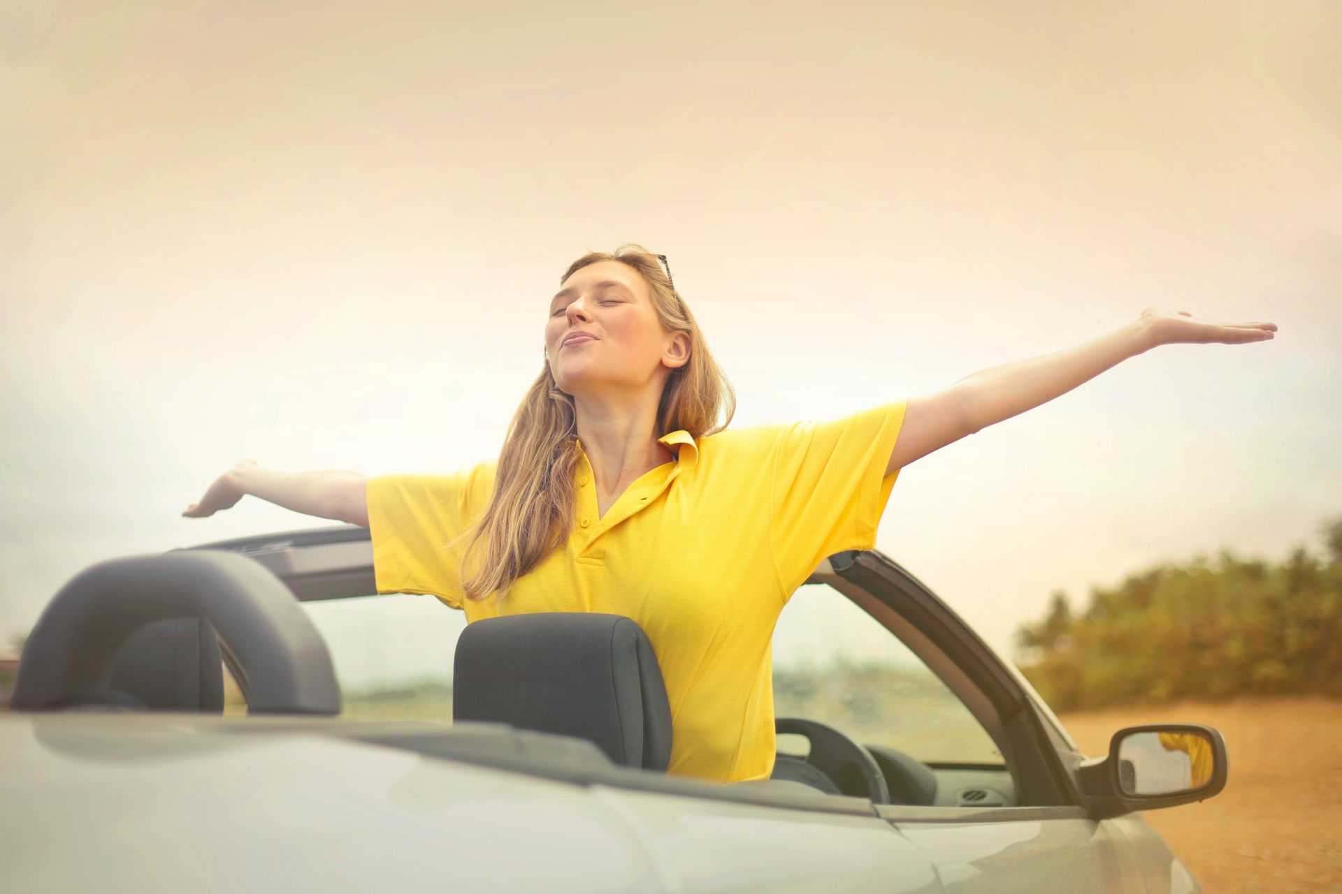 Woman in yellow shirt, arms outstretched in a convertible car, enjoying the sun.
