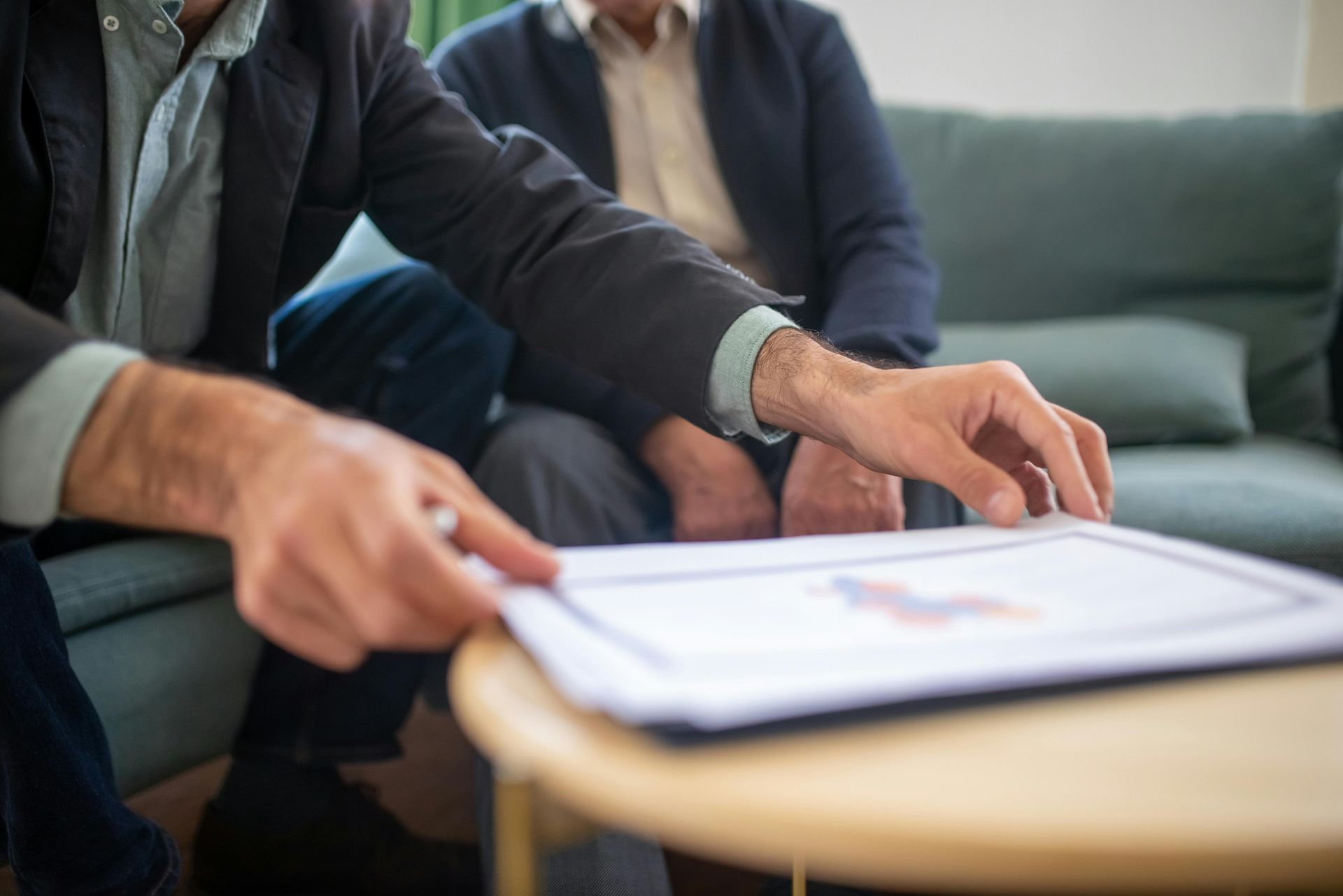 Person in blazer pointing at a document on a table, with another person sitting on a couch.
