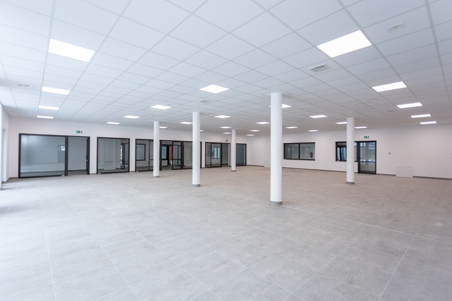Empty, open office space with columns, recessed lighting, gray tile floor, and glass-paneled entryways.