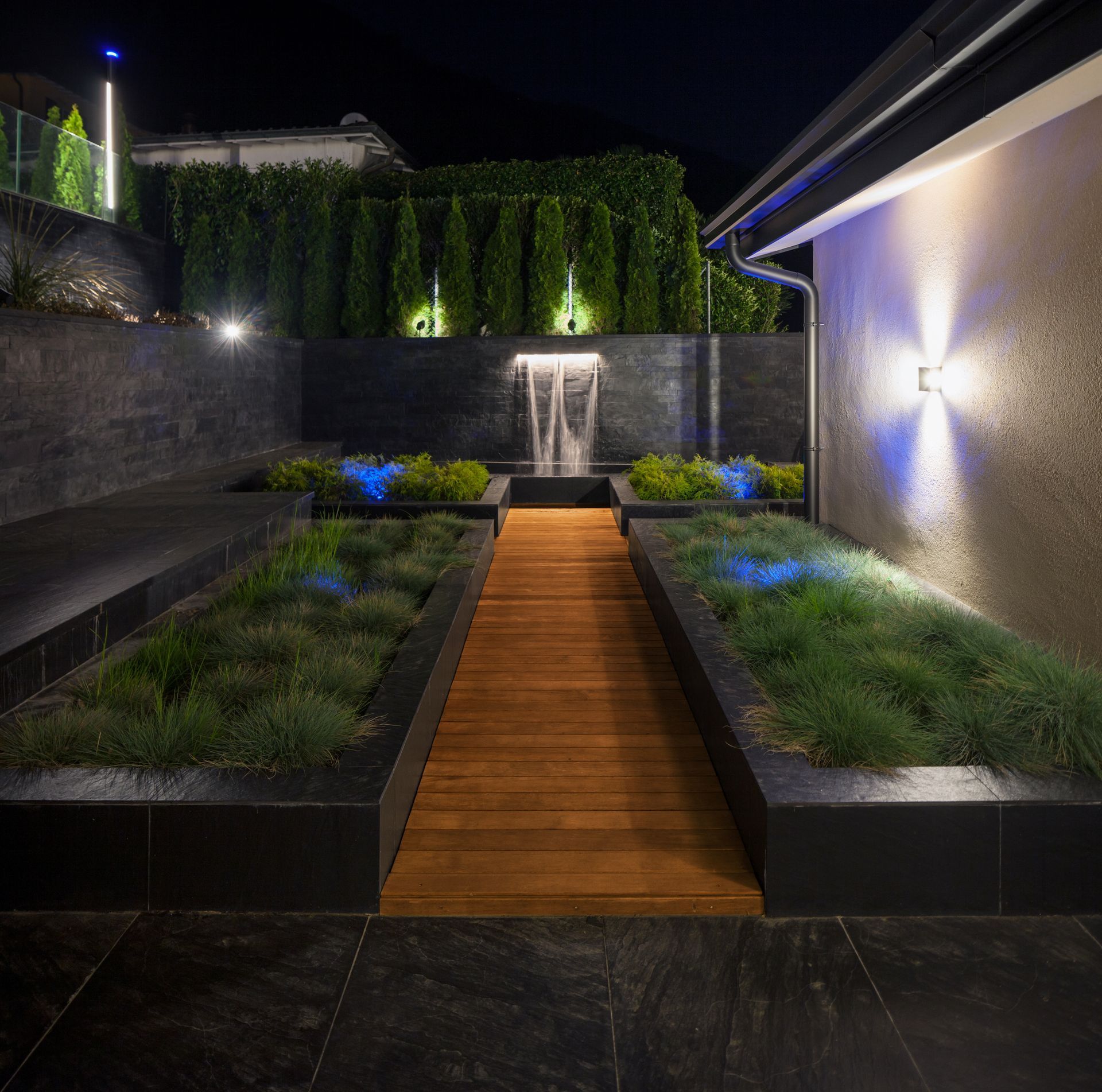 Night view of a modern garden with a wooden walkway, water feature, and illuminated greenery.