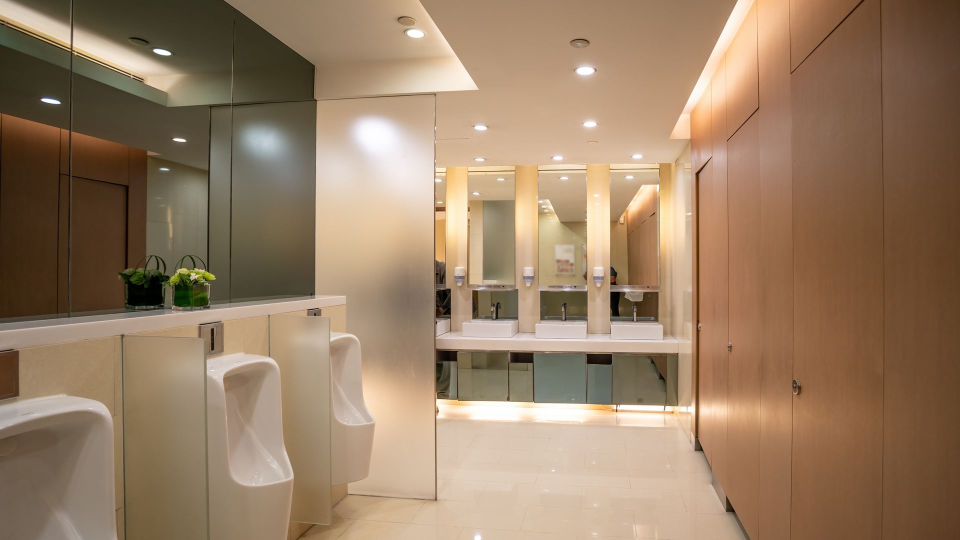 Public restroom with urinals, sinks, mirrors, and stalls; light wood and white tile, modern design.