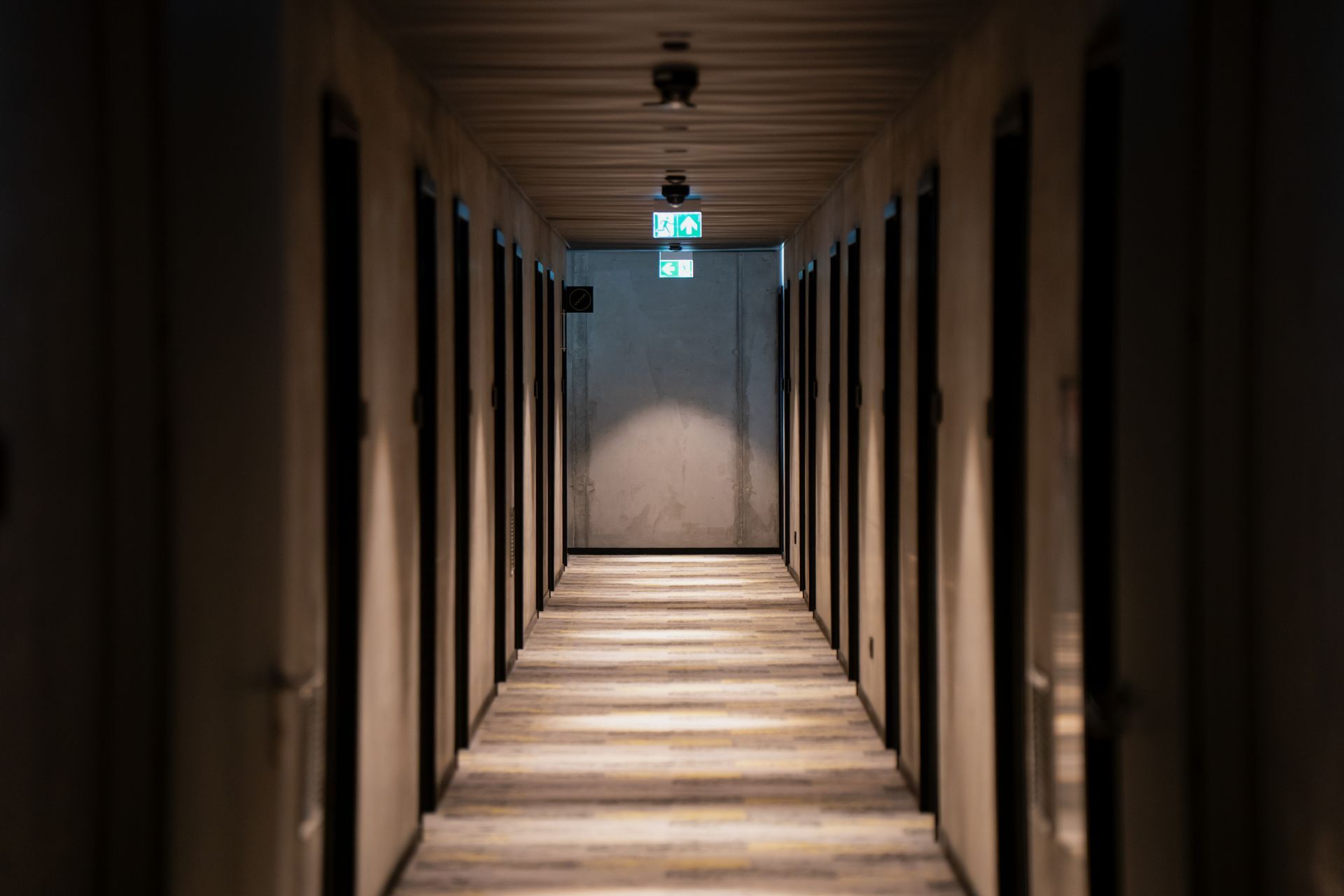 Long, dimly lit hotel hallway with doors on both sides, leading to an emergency exit sign at the end.