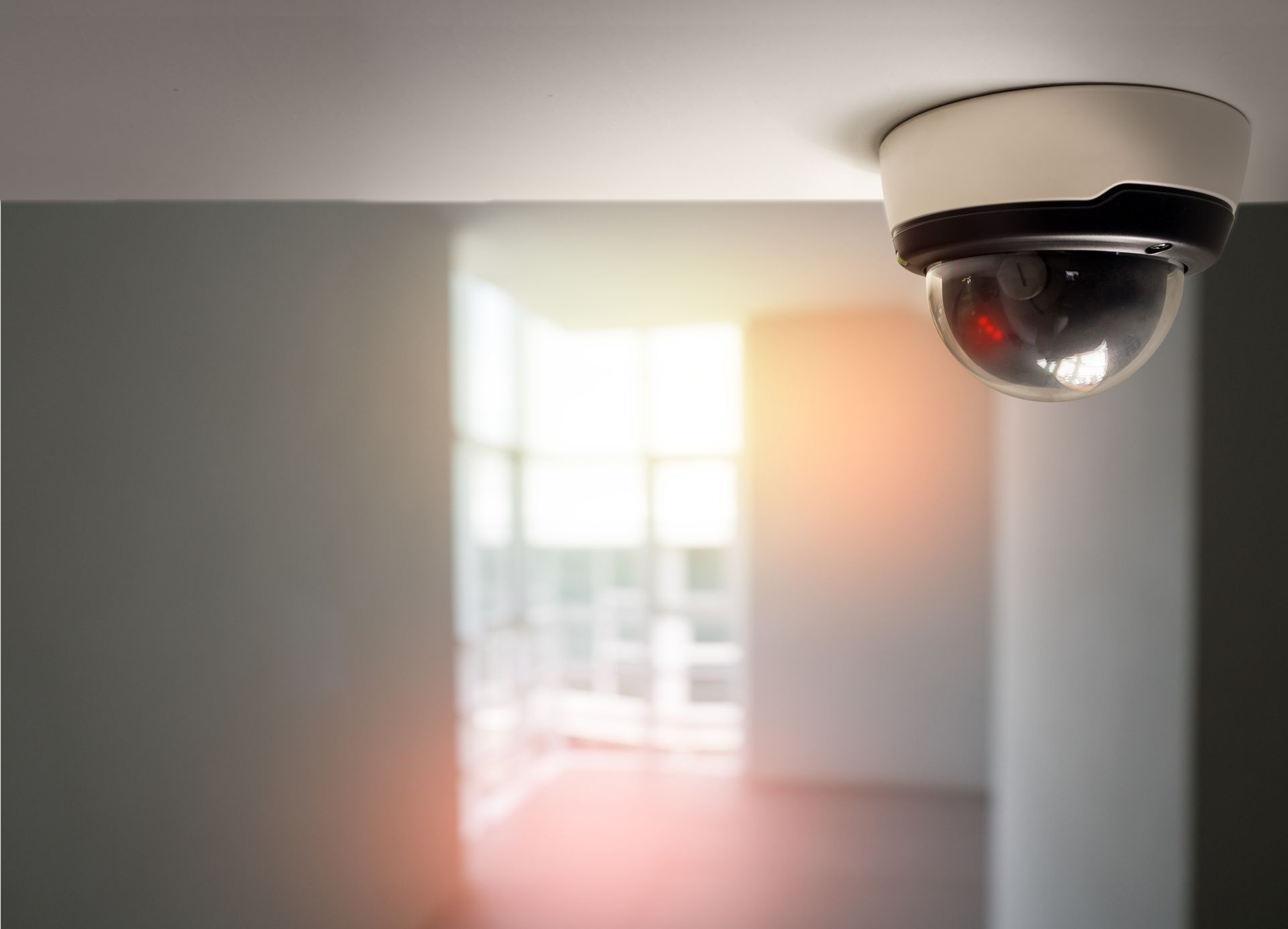 Security camera mounted on a white ceiling, overlooking an empty room with bright windows.