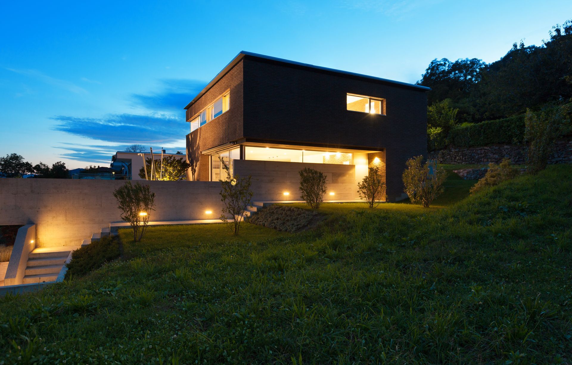 Modern two-story house with dark facade, illuminated windows, and surrounding lawn at dusk.