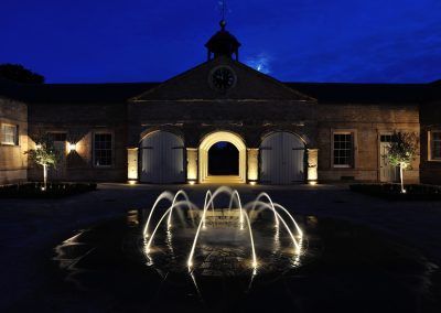 Night view of a fountain with arched water jets in front of a building with arched entryways and a clock tower.