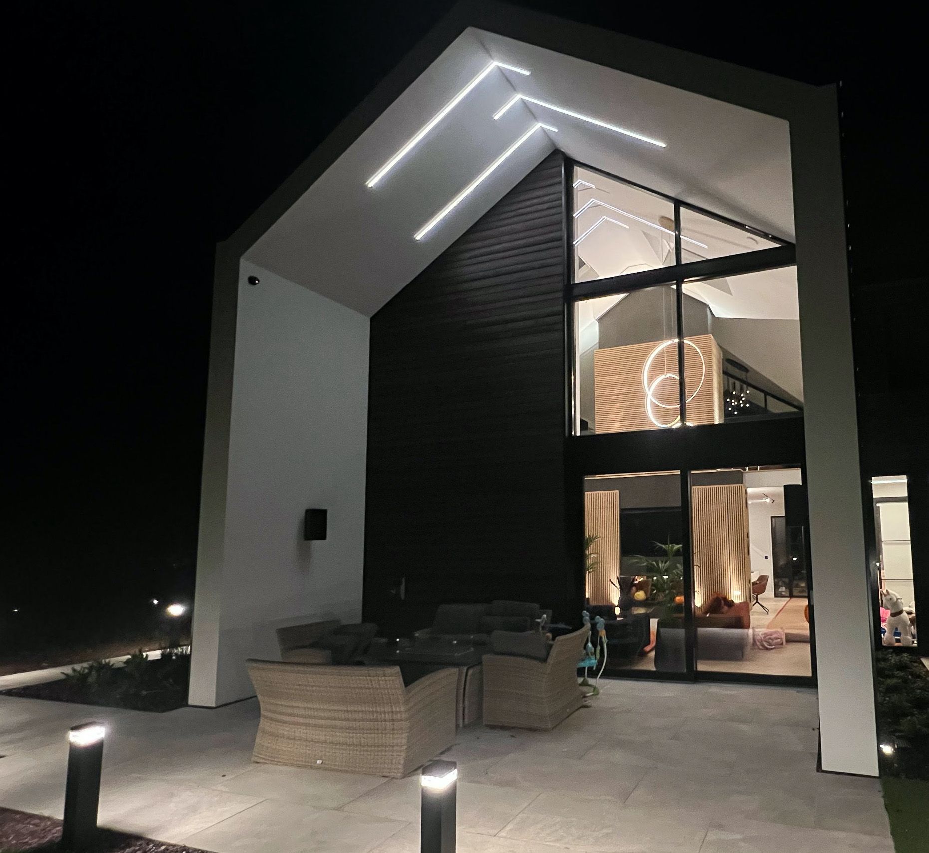 Modern house exterior at night, with illuminated roof, large windows, and patio seating.