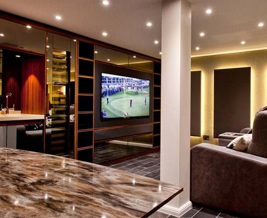 Luxury basement media room with TV, shelving, bar, and seating. Dark wood accents, recessed lighting.