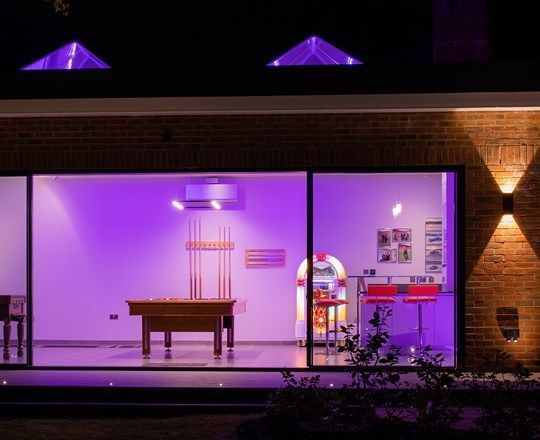 Exterior view of a game room lit with purple lights, featuring a pool table, jukebox, and bar.