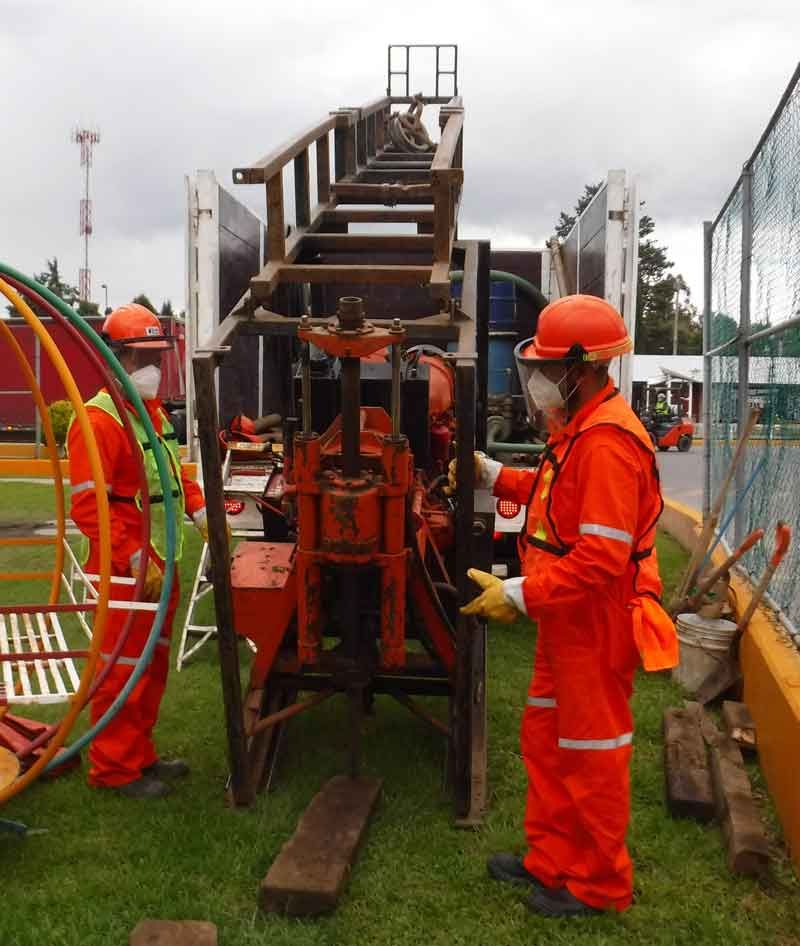 Dos trabajadores con trajes protectores de color naranja operan una máquina perforadora en una zona cubierta de césped cerca de un parque infantil.