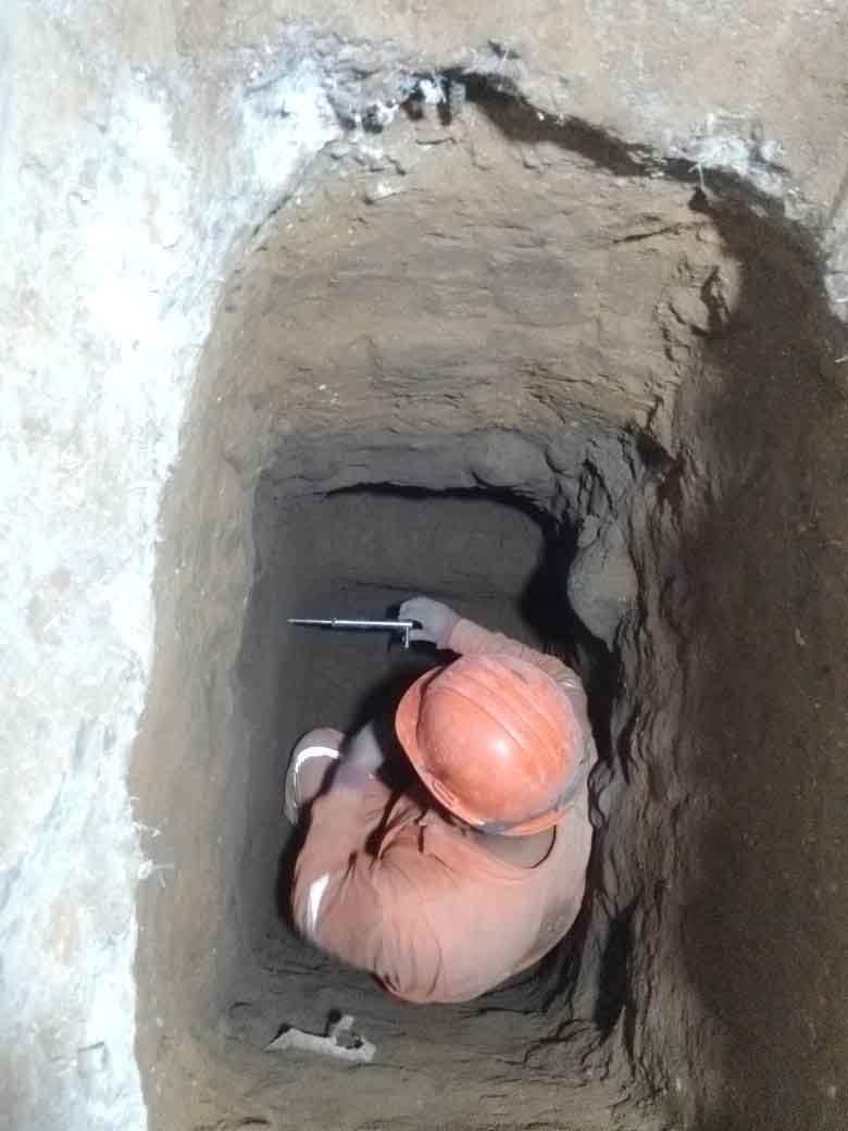 Persona con ropa de trabajo naranja y casco en un pozo profundo y estrecho. Sostiene una herramienta y trabaja.