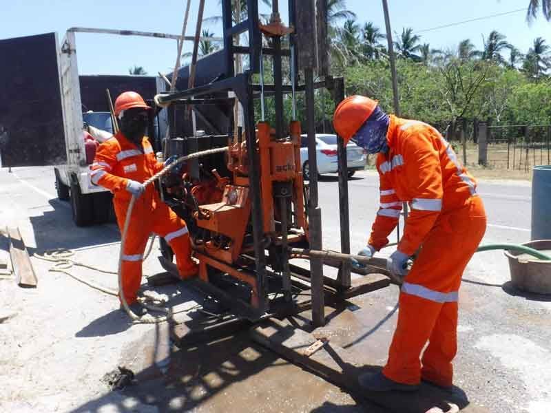 Dos trabajadores con monos de color naranja operan equipos de perforación al aire libre cerca de una carretera.