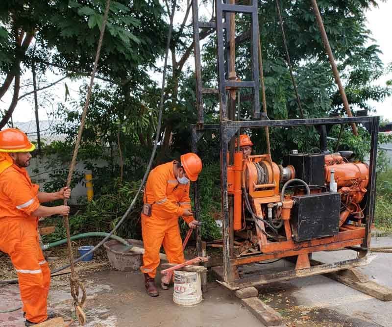 Trabajadores con overoles de color naranja operan una plataforma de perforación cerca de árboles, probablemente para realizar pruebas de suelo.