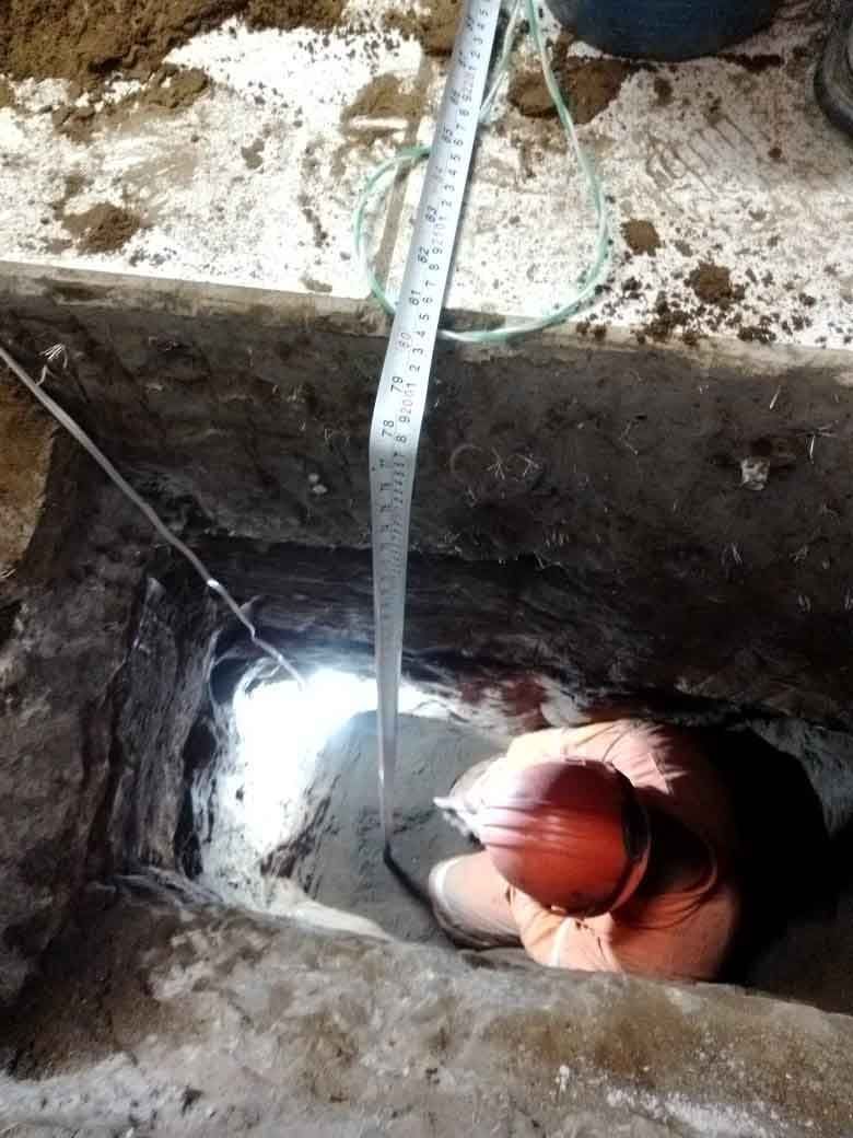 Una persona con casco mide la profundidad de un agujero con una cinta métrica. El agujero está en una zona polvorienta y de color claro.