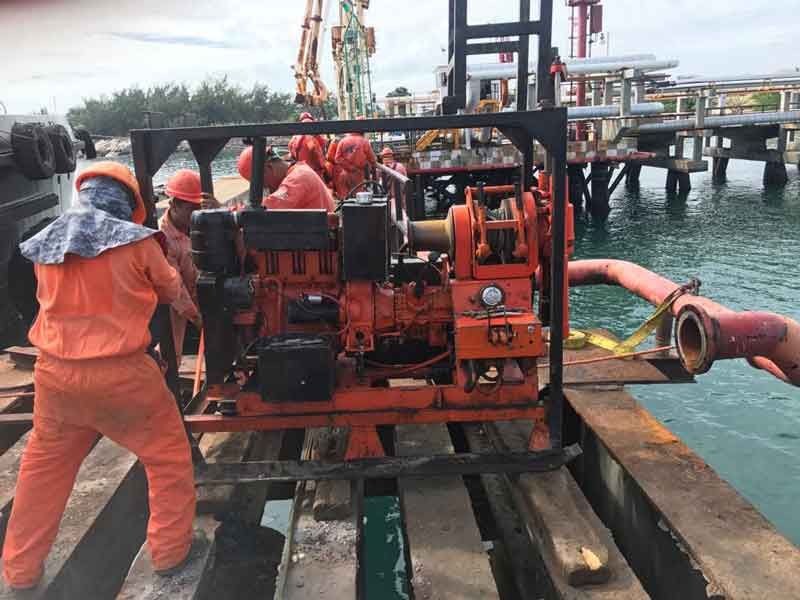 Trabajadores con trajes naranjas mueven una bomba naranja en un muelle cerca del agua.