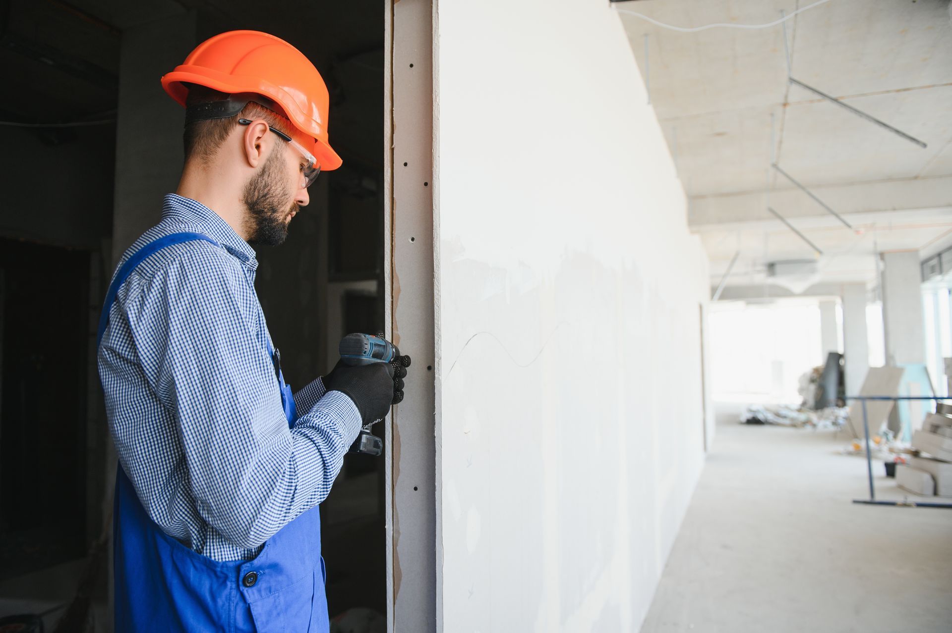 Construction worker in orange hard hat and blue overalls using a drill on a wall in a building under construction.